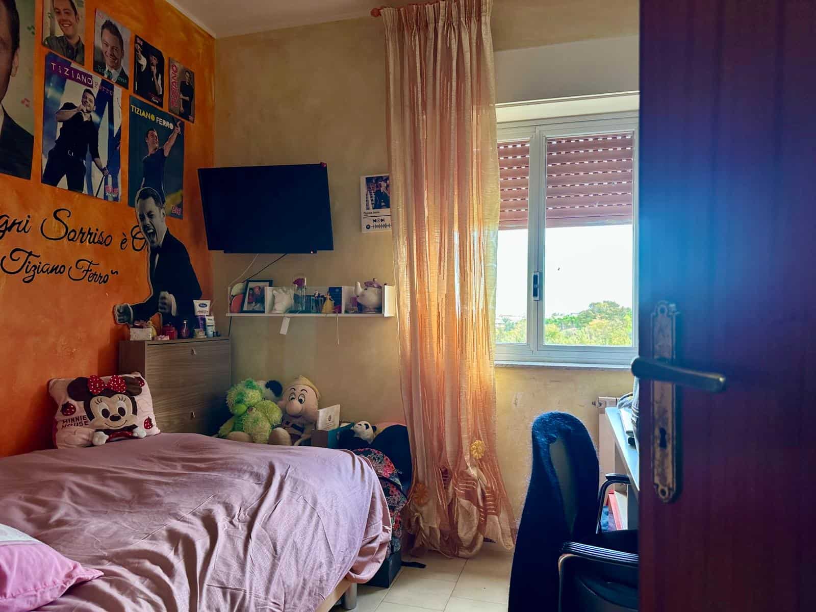 1. Stanza da letto con decorazioni di personaggi Disney e poster politici, letto con copripiumino rosa, soggiorno luminoso con finestra e tende arancioni, ambiente accogliente e personalizzato.