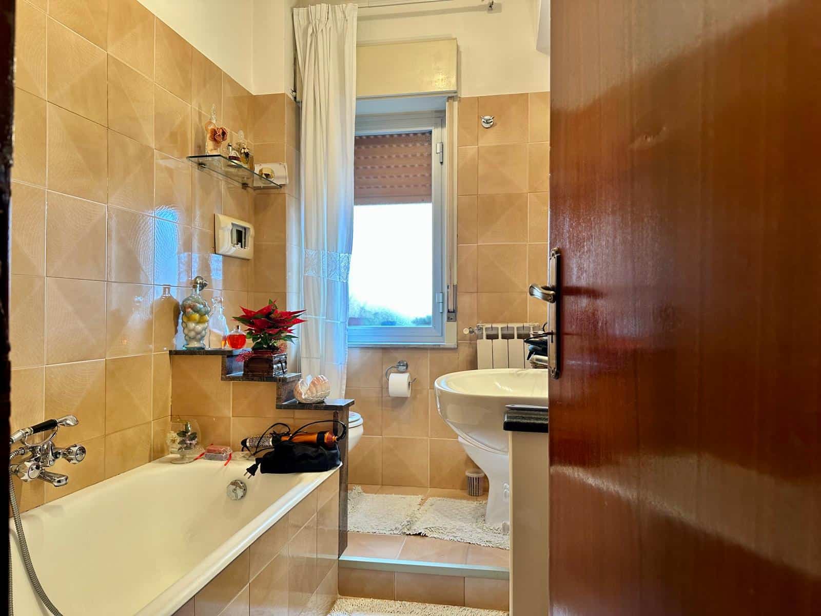 Bagno accogliente con vasca, lavabo e servizi essenziali, pareti rivestite di piastrelle beige, finestra naturale, decori natalizi e oggetti di arredamento, ideale per una casa moderna e funzionale.
