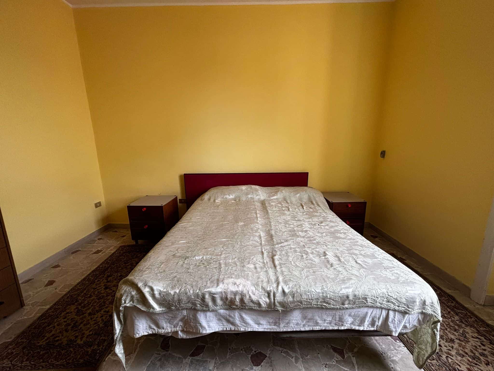 Elegante camera matrimoniale con pareti gialle, letto con lenzuola brillanti e comodini in legno, perfetta per affitti turistici e residenziali, in zona tranquilla.