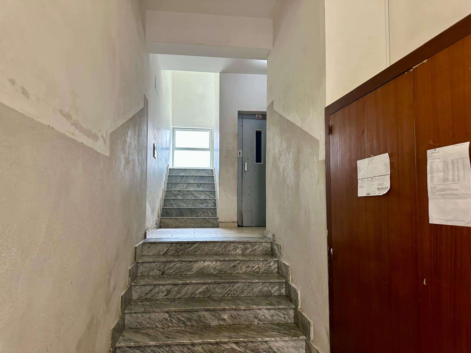 Ascensore e scale nel condominio, ingresso in palazzina con ampie finestre e pareti in declino, ambienti interni con finiture in marmo.