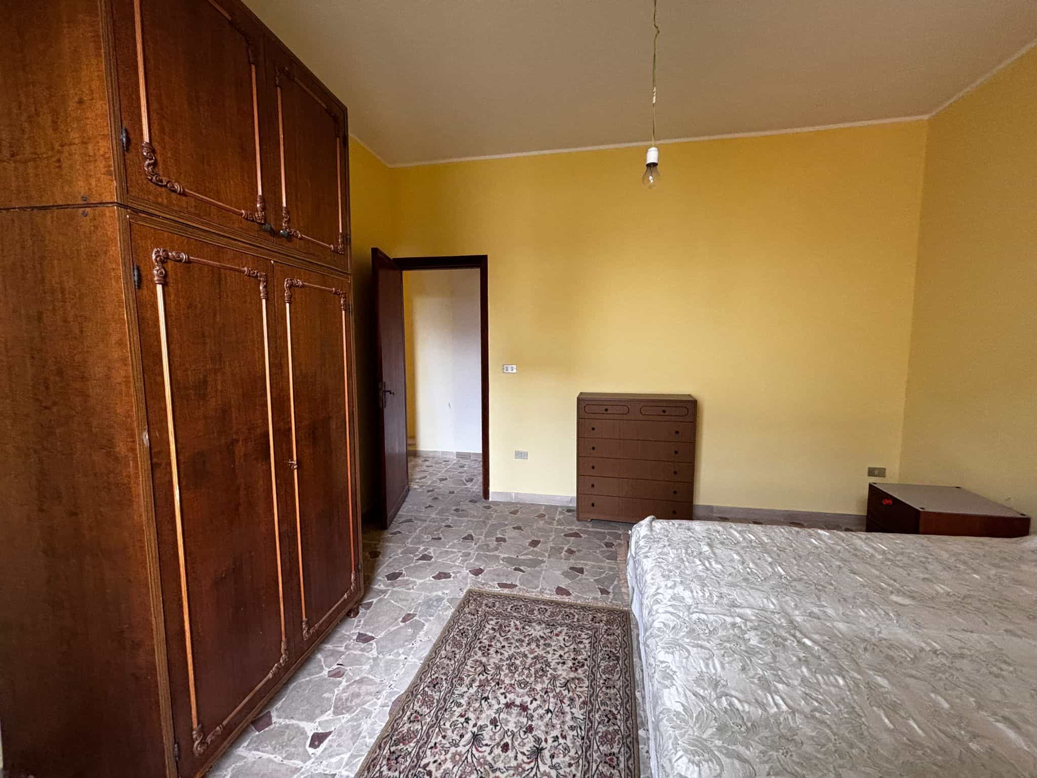 Armadio in legno massiccio in camera da letto con pareti gialle, pavimento in pietra, letto matrimoniale e comò vintage, arredamento classico e un ambiente spazioso e luminoso.