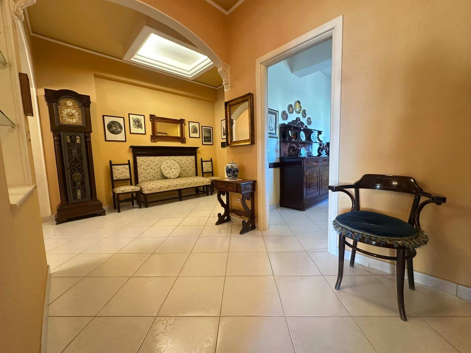 Antico salotto arredato con mobili in vero legno, tra cui orologio a colonna, quadro d'autore e credenza con porcellane, con pareti color ocra e soffitto a cassettoni.