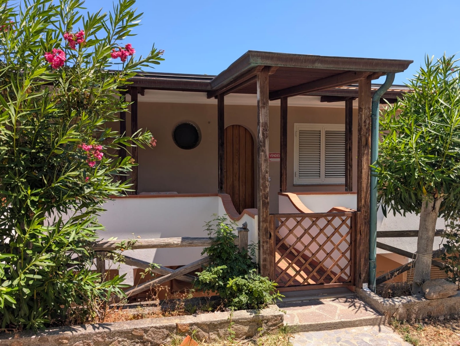 Balcone in legno con porta arcuata, circondato da piante verdi e fiori rosa, in una casa in stile tradizionale vicino al mare, perfetta per vivere in tranquillità e relax.