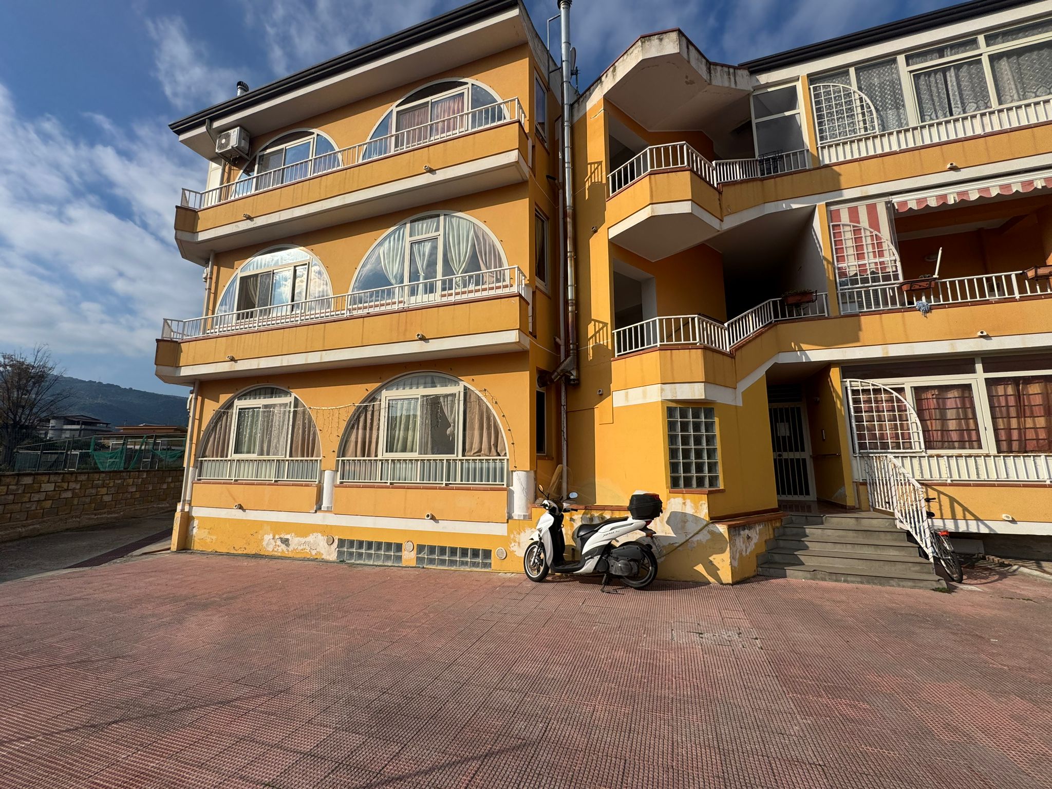 Ampio appartamento in stabile con balconi, facciata gialla e accesso con scale, parcheggio esterno, motorino e biciclette, ideale per investimento immobiliare a NextCasa.