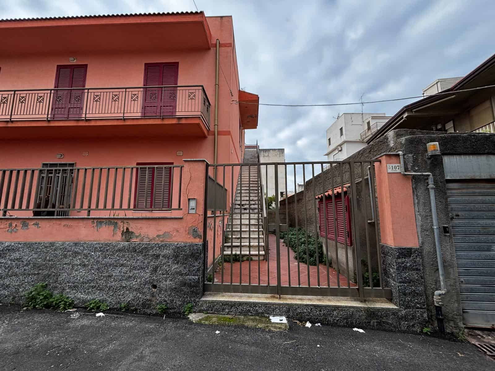 Finestra e balcone di una casa residenziale in stile italiano, mimetizzata in un quartiere urbano, con sguardo sulla strada e un cielo nuvoloso, perfetta come immagine per annunci immobiliari e intermediazioni.