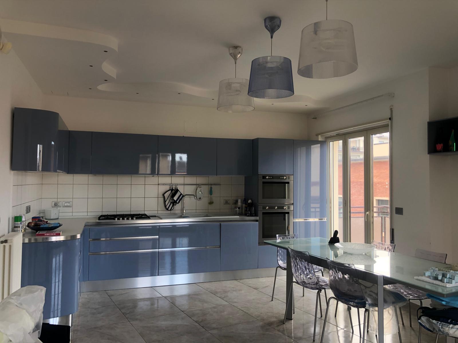 Lavastoviglie moderna in cucina con mobili blu, tavolo in vetro e sedie trasparenti, illuminazione a soffitto e grande finestra con accesso al balcone, ambiente luminoso e elegante.