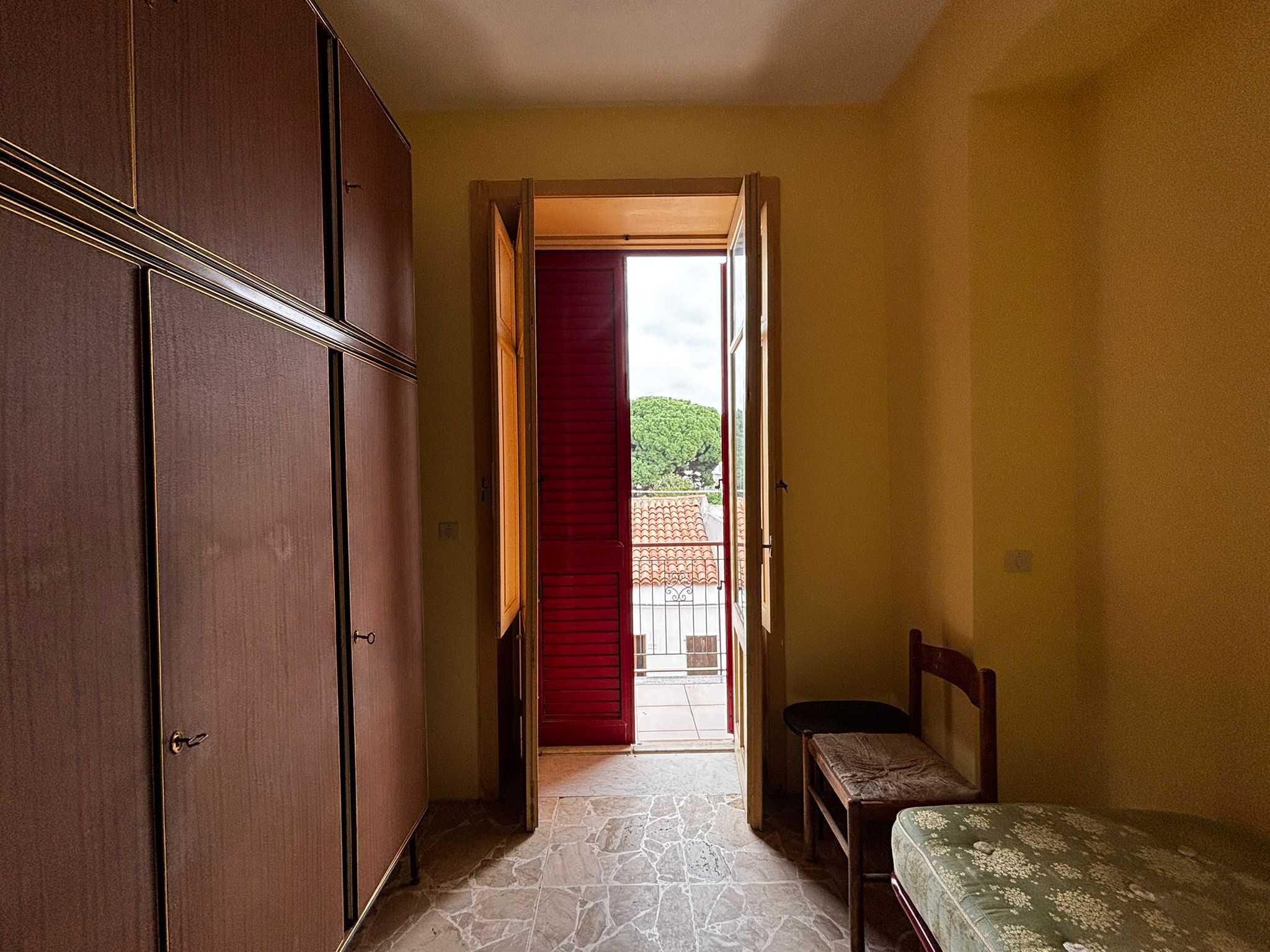 Spetto di camera da letto con armadio in legno, finestra con persiana rossa, vista su tetti e alberi, interni accoglienti, ideale per affitti immobiliari, NextCasa.