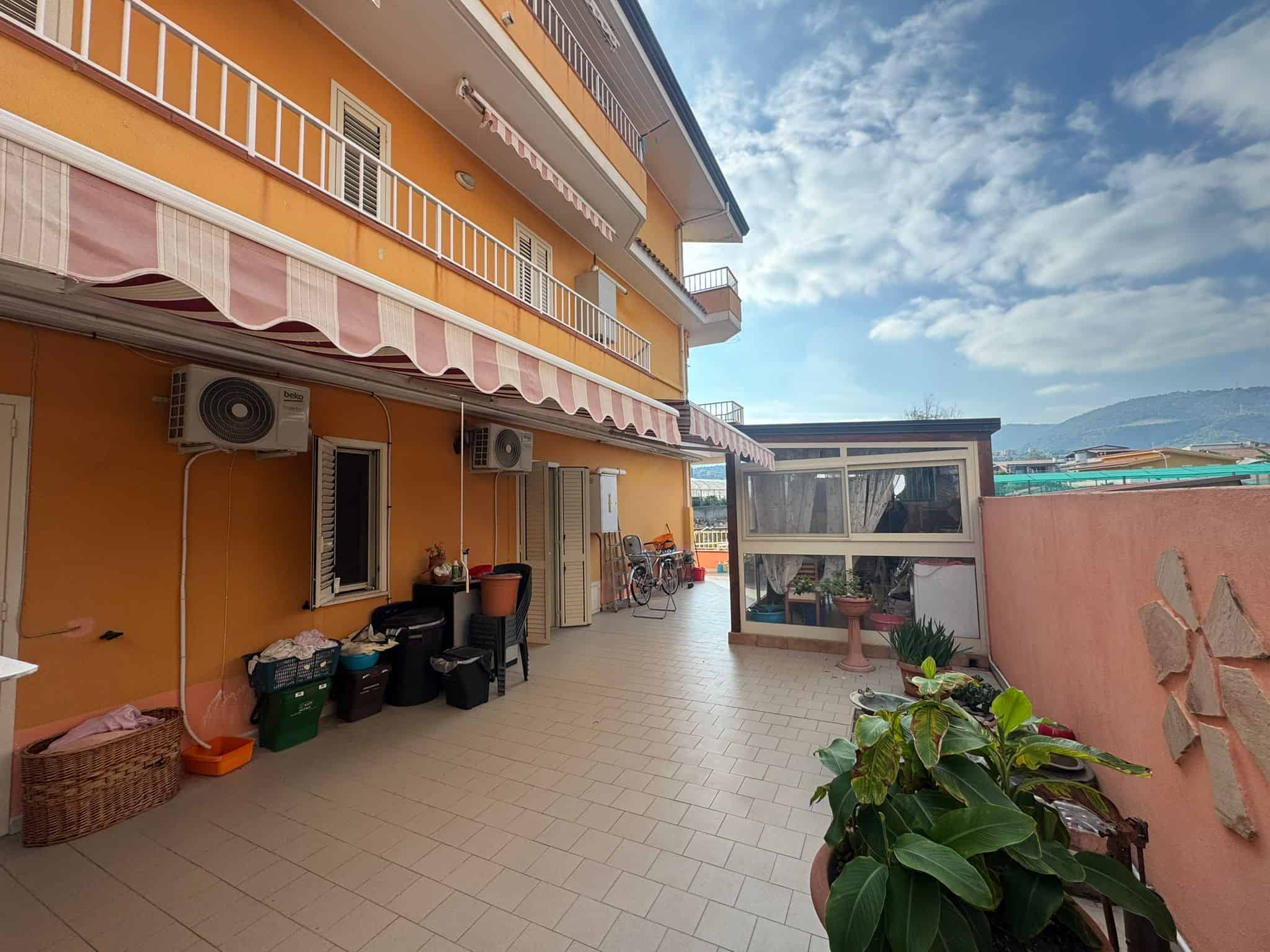 Ampio terrazzo con vista sul mare e montagne, ideali per vivere e rilassarsi all'aperto in residence di Napoli. Spazio pratico e accogliente con piante, arredamento funzionale, e tende oscuranti.