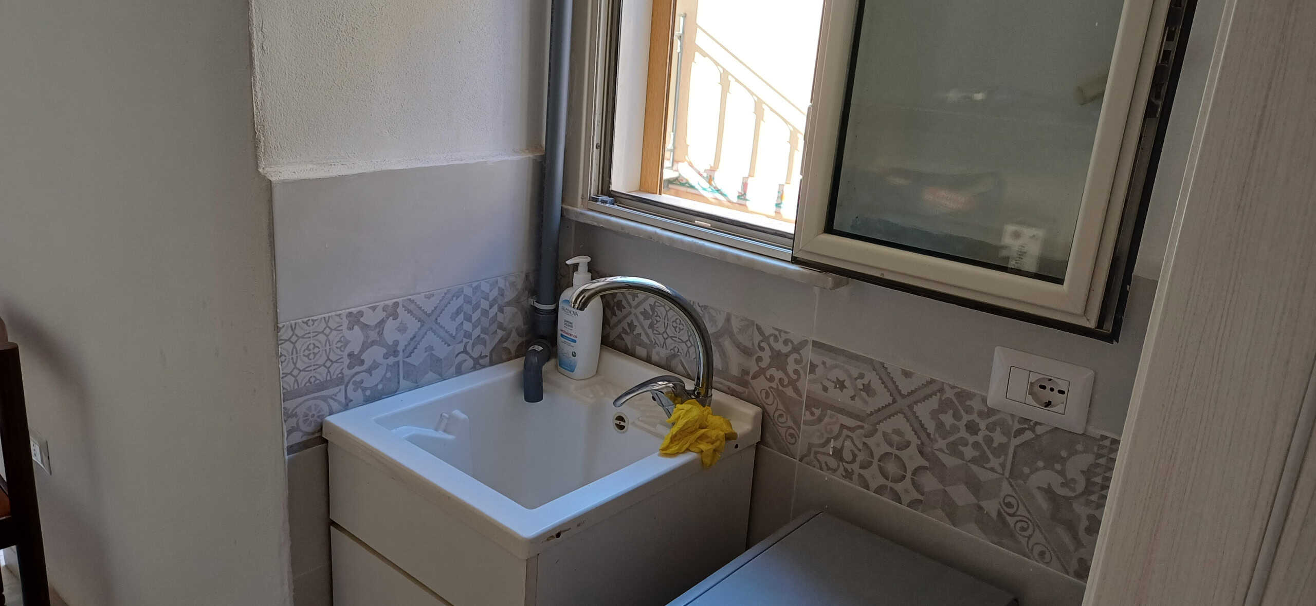 Lavandino da appartamento in zona lavanderia, vicino alla finestra con vista sulla balaustra. Stanza funzionale con rivestimenti decorativi e presa di corrente.