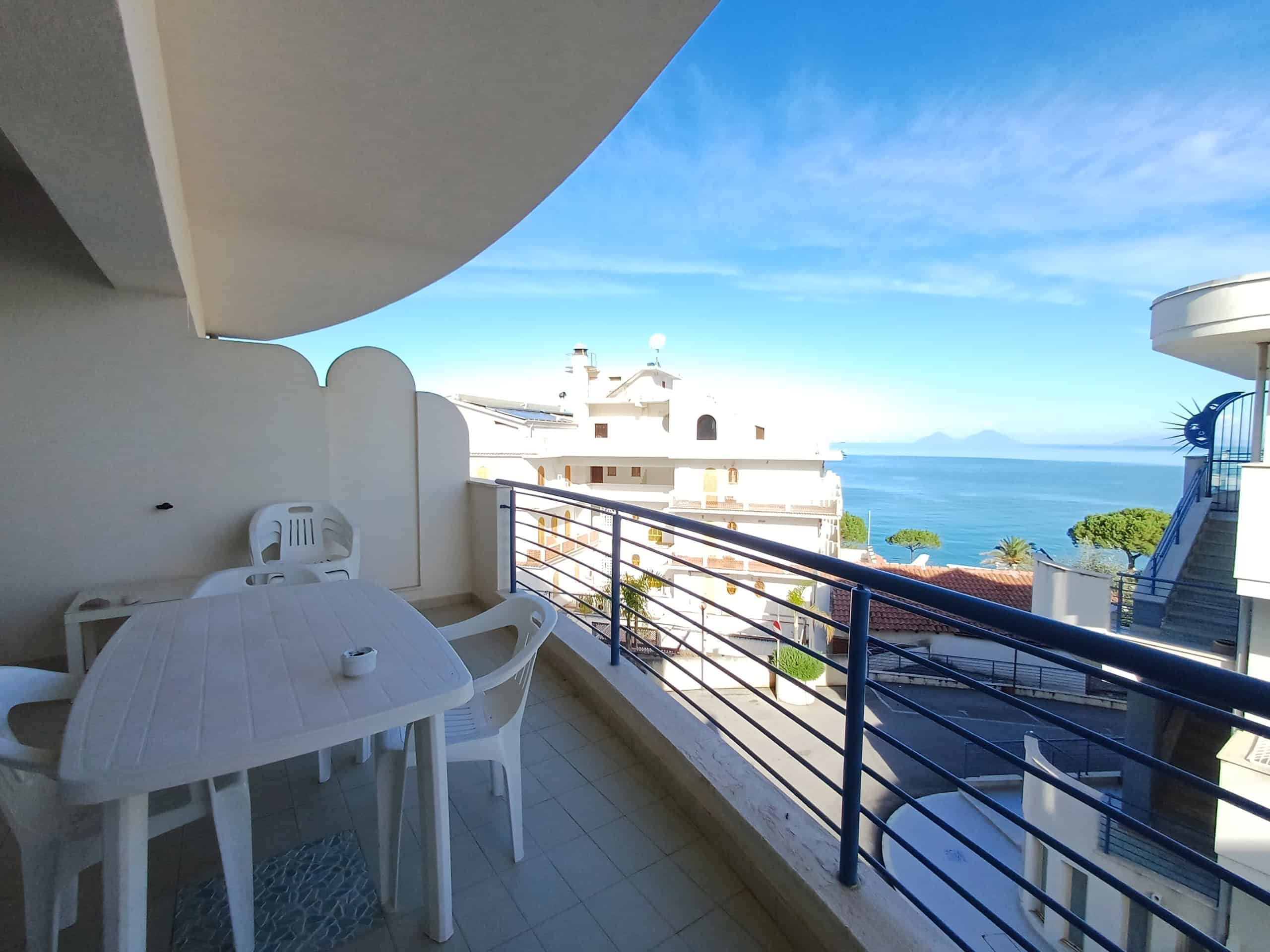 Vista panoramica sul mare dal balcone di un appartamento in affitto, con tavolo in plastica e sedie, in zona residenziale con edificio moderno e cieli azzurri, ideale per vacanze e relax.