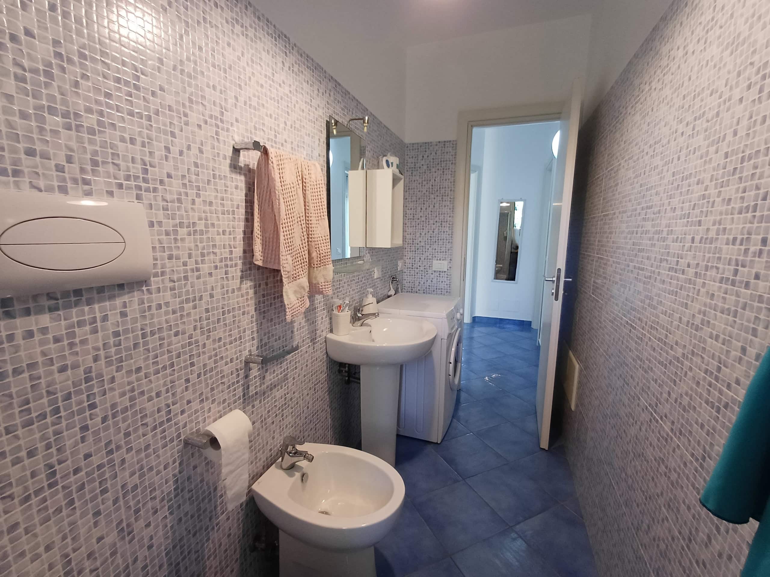 Bagno moderno con mosaico piastrelle azzurro, lavabo, bidet, asciugamani rosa, armadietto bianco, pavimento blu, luce naturale, spazio funzionale e ben arredato.