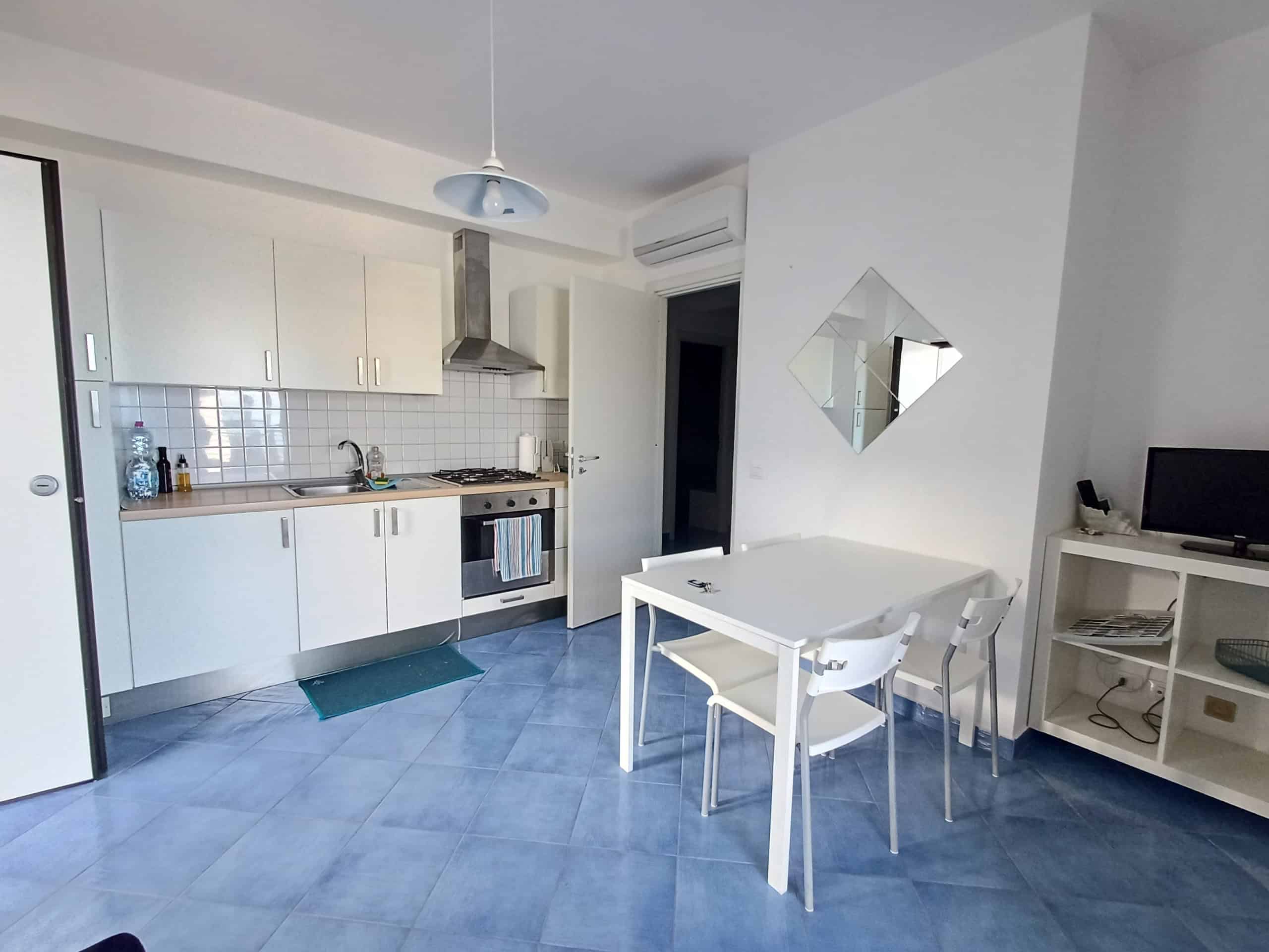 Appartamento moderno con cucina attrezzata, soggiorno luminoso, area pranzo con tavolo e sedie, arredo minimalista, parete con specchio decorativo, TV su mobile, ambiente accogliente e funzionale.