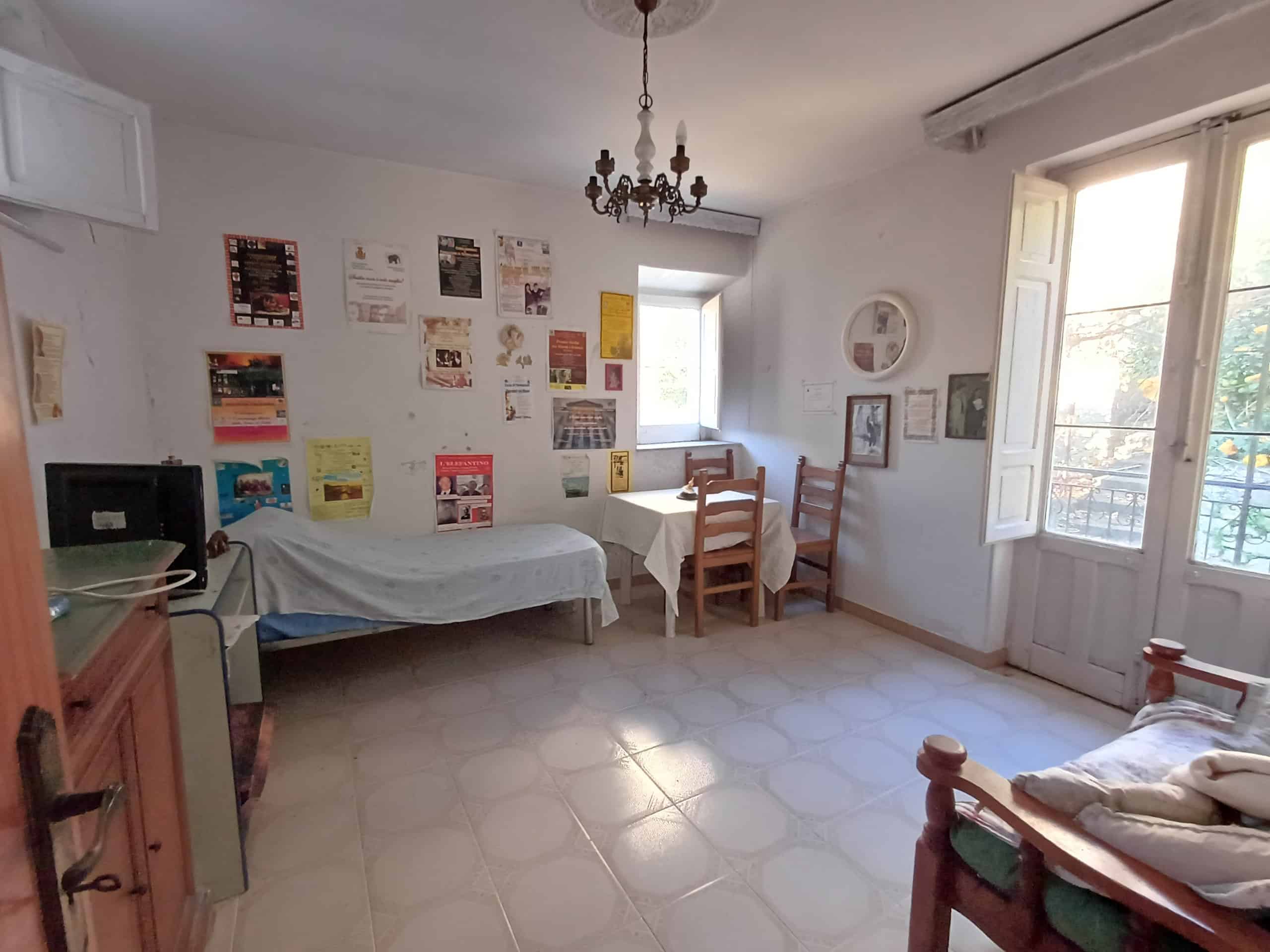 Ampia stanza rustica con tavolo, sedie, divano e mobili in legno, illuminata da luce naturale, decorata con poster e quadri, ideale per soggiorno o sala da pranzo in casa tradizionale italiana.
