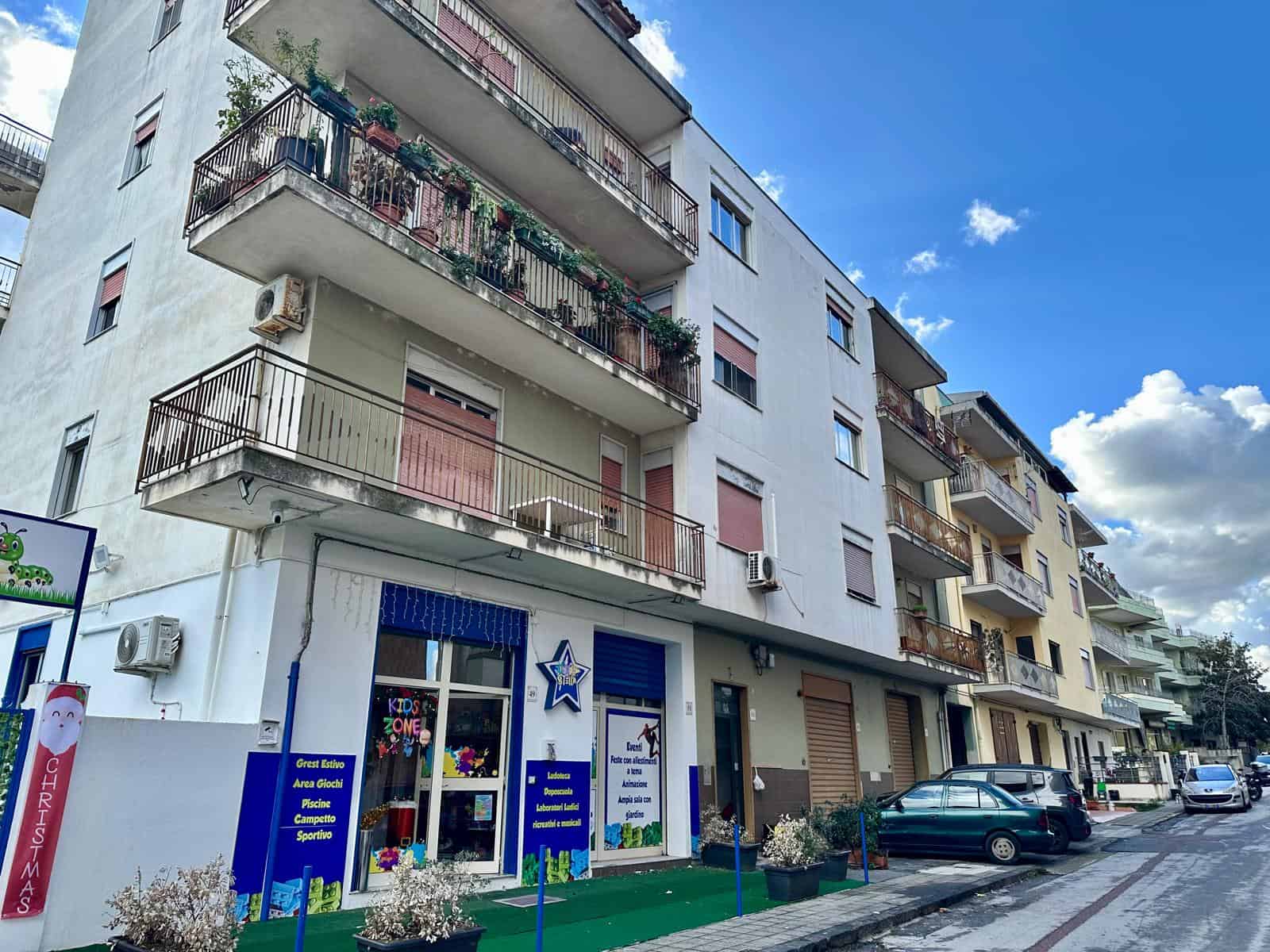 Balcone di un appartamento in edificio residenziale con piante e fiori, su una strada urbana con negozio di giochi e parcheggio, cielo sereno e nuvole bianche, atmosfera di quartiere.