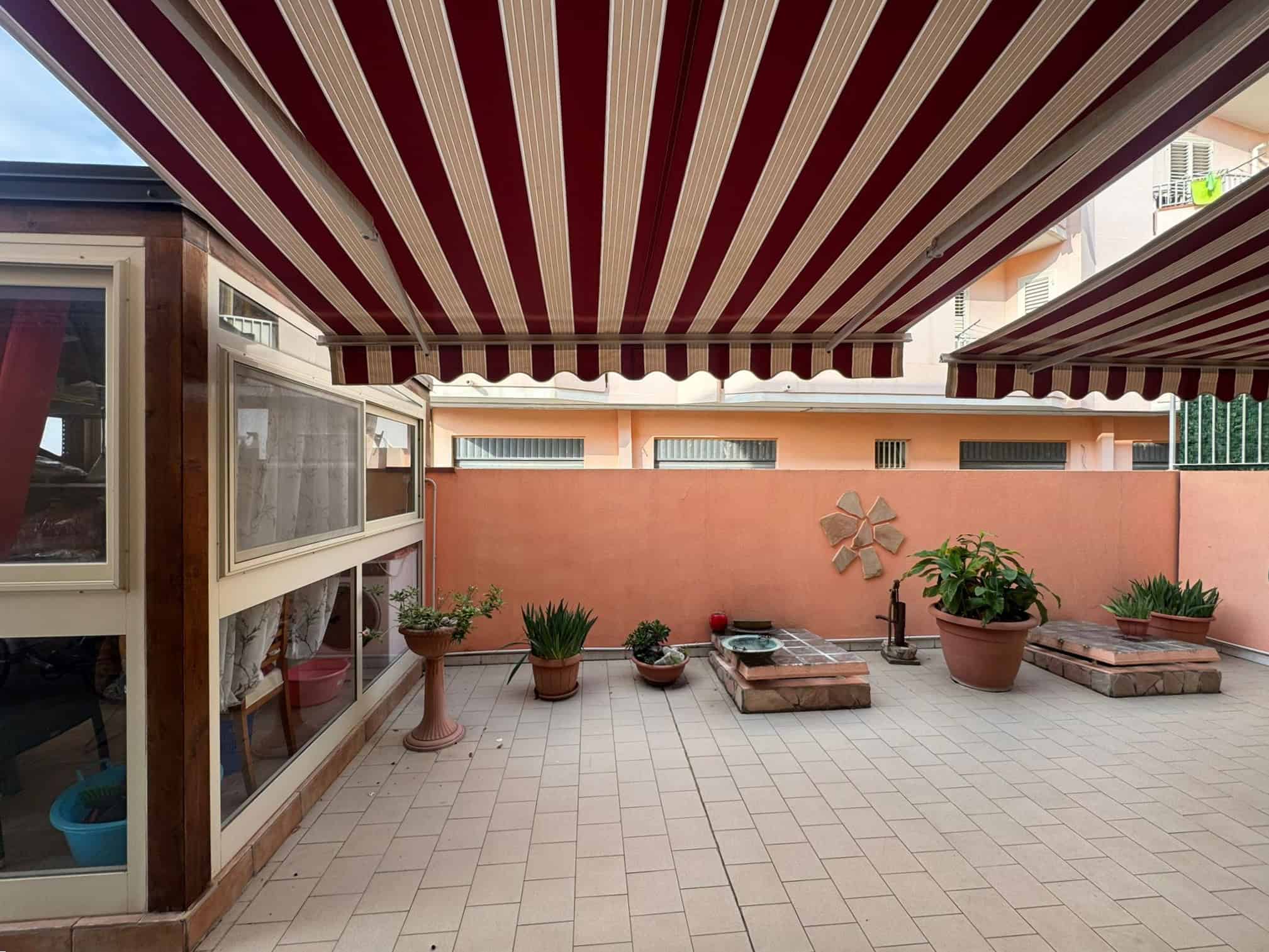 Ampio terrazzo con tenda a strisce rosse e beige, piante in vasi decorativi, angolo relax e spazio, zona esterna ideale per vivere all'aperto.