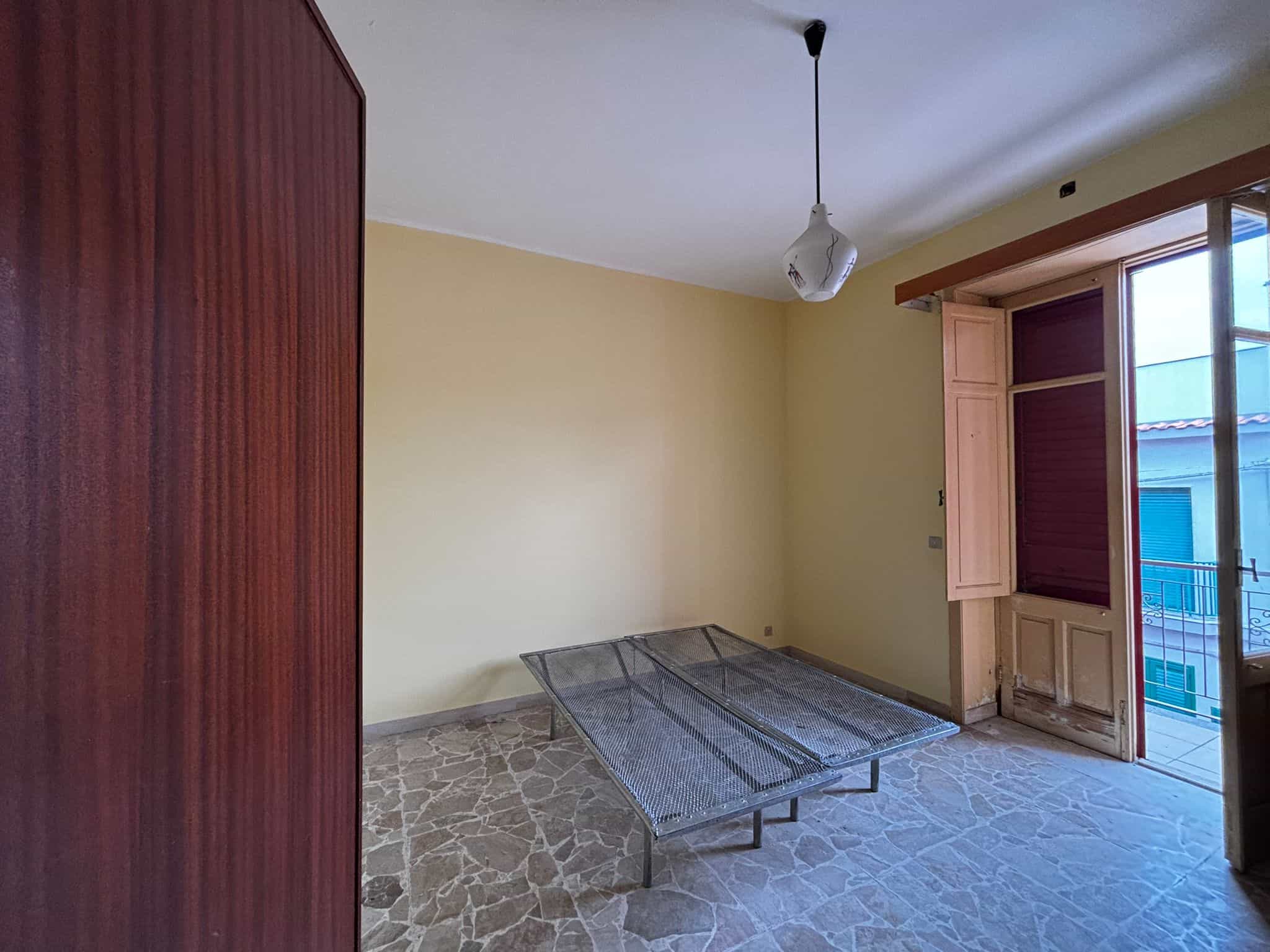 Soggiorno vuoto con pareti gialle, grandi finestre con infissi in legno e balcone, arredamento minimal, pavimento in pietra, luce naturale, ideale per investimento immobiliare.