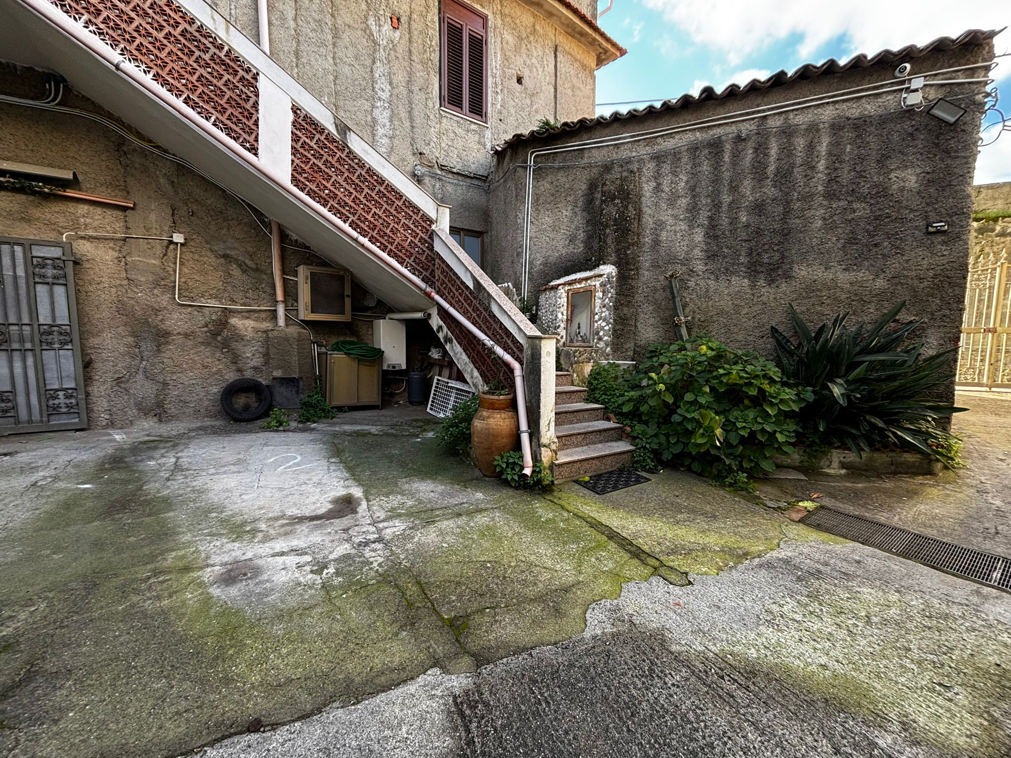 Vecchio cortile esterno con scale e piante, edificio residenziale con pareti in pietra e cemento, atmosfera urbana, stile semplice, con elementi di recupero, illuminazione naturale.