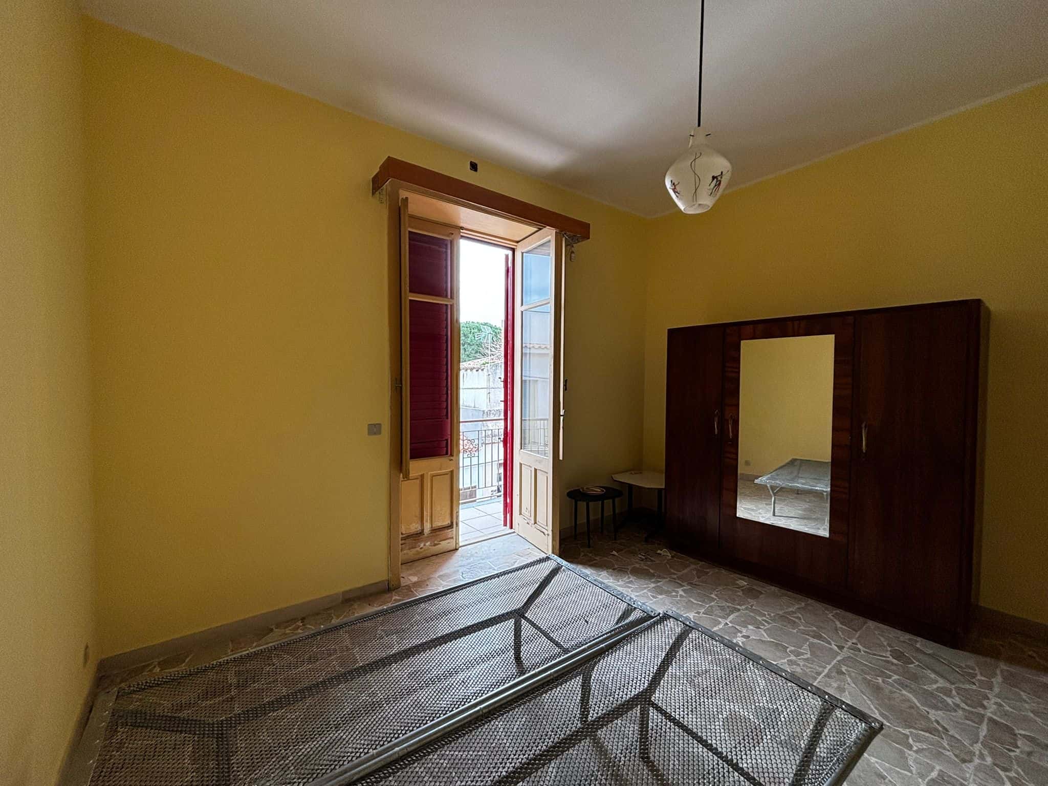 Letto singolo in camera da letto arredata, stanza luminosa con porta balcone, armadio in legno, pareti gialle, pavimento in pietra, ambiente confortevole e tranquillo, ideale per affitti a breve o lungo termine.