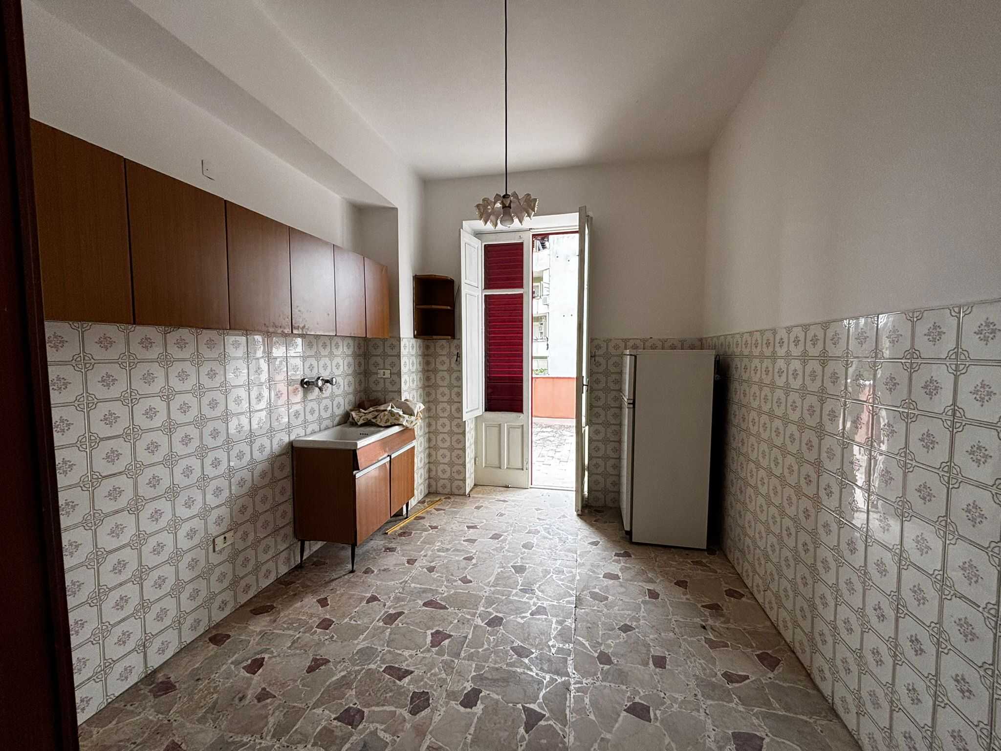 Vecchia cucina con piastrelle decorative, armadietti in legno, frigorifero e accesso a un balcone soleggiato, ideale per ristrutturazione o investimento immobiliare.