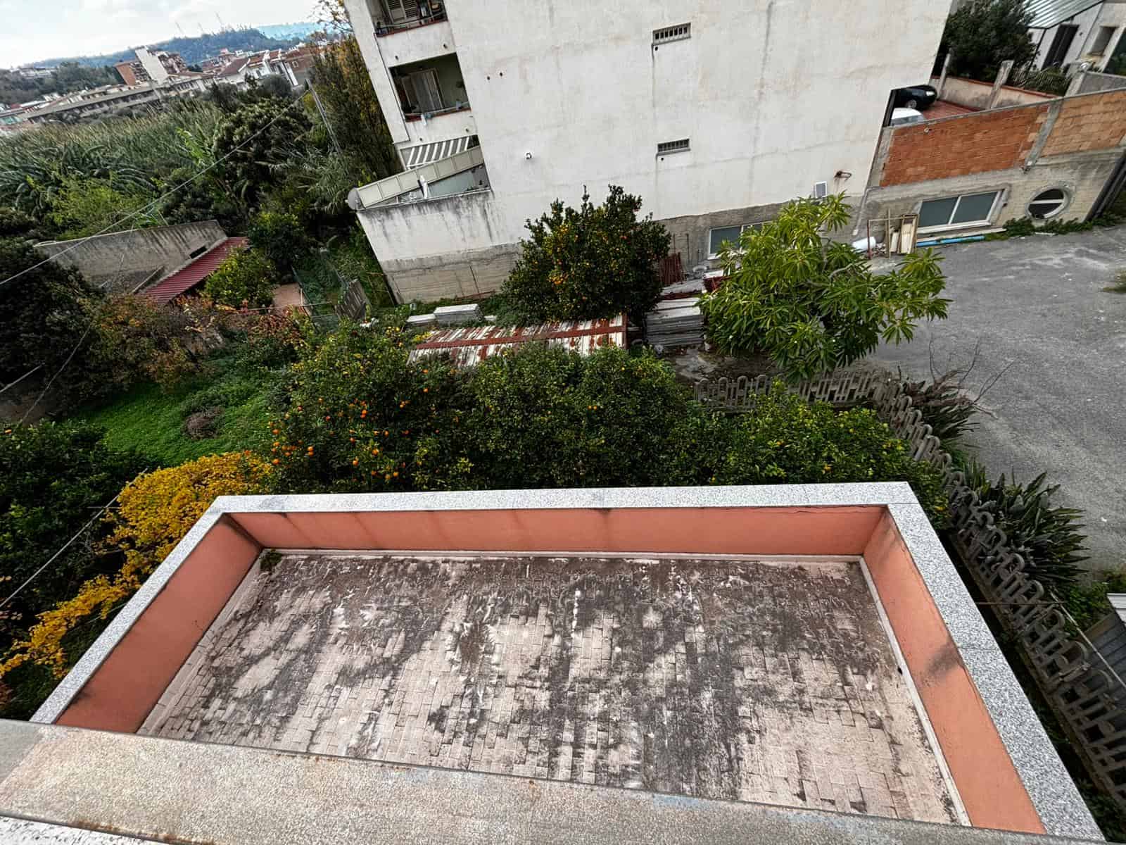 Balcone di un appartamento con vista su giardino con alberi e piante, in un complesso residenziale urbano, con edifici vicini e spazio esterno.