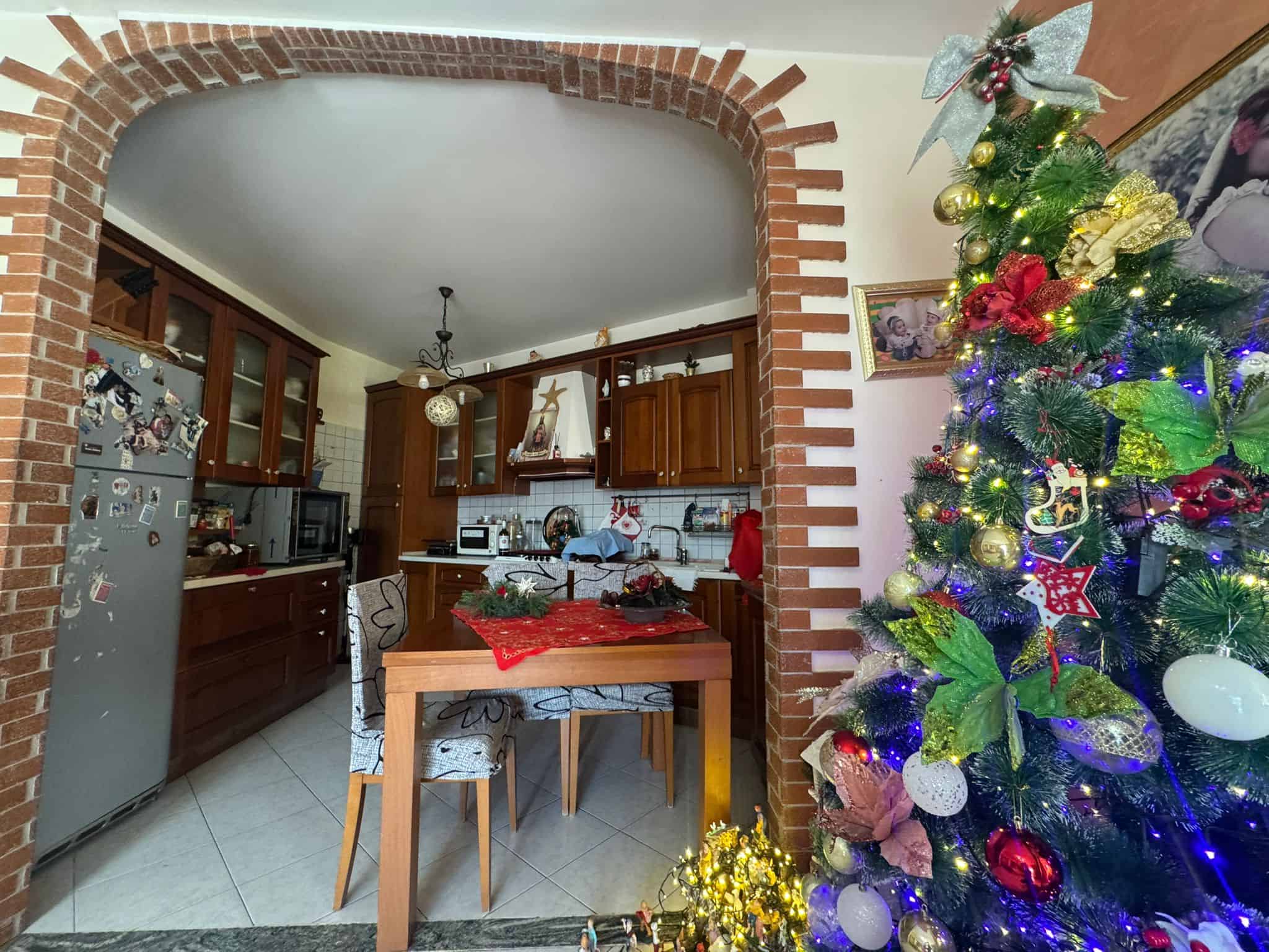Acciaio di Natale decorato con luci e ornamenti colorati, ambientazione festiva in cucina con arco in mattoni rossi, tavolo in legno e dettagli natalizi.