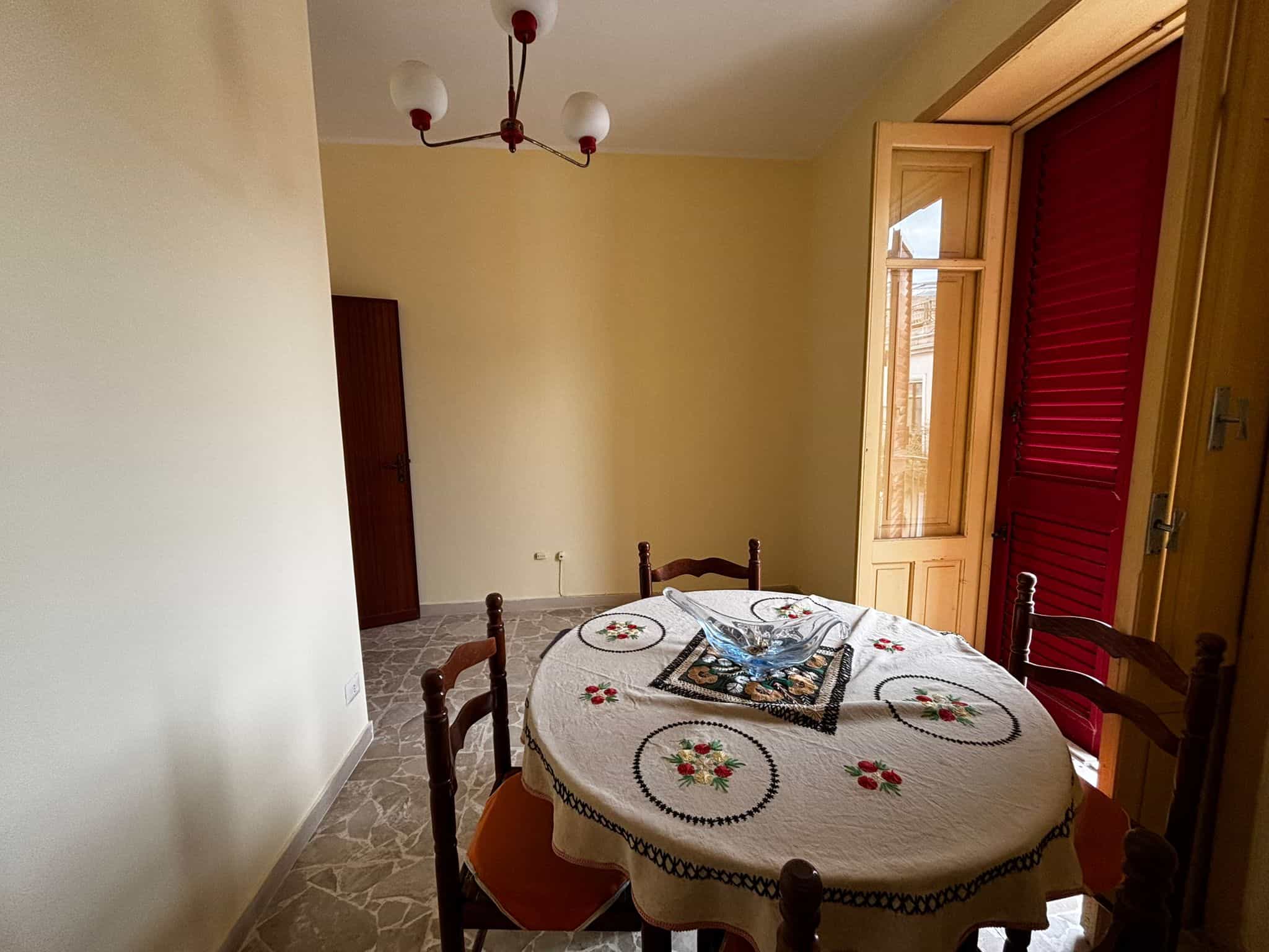 1. Angolo pranzo con tavolo rotondo, sedie in legno e vista su finestra con persiane rosse e apertura su balcone, illuminato dalla luce naturale.