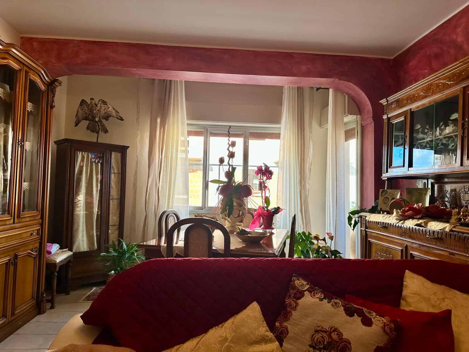 1. Interior soggiorno con mobili in stile classico, divano rosso con cuscini decorativi, tavolo da pranzo vicino alla finestra e lampade, ambiente caldo e accogliente.