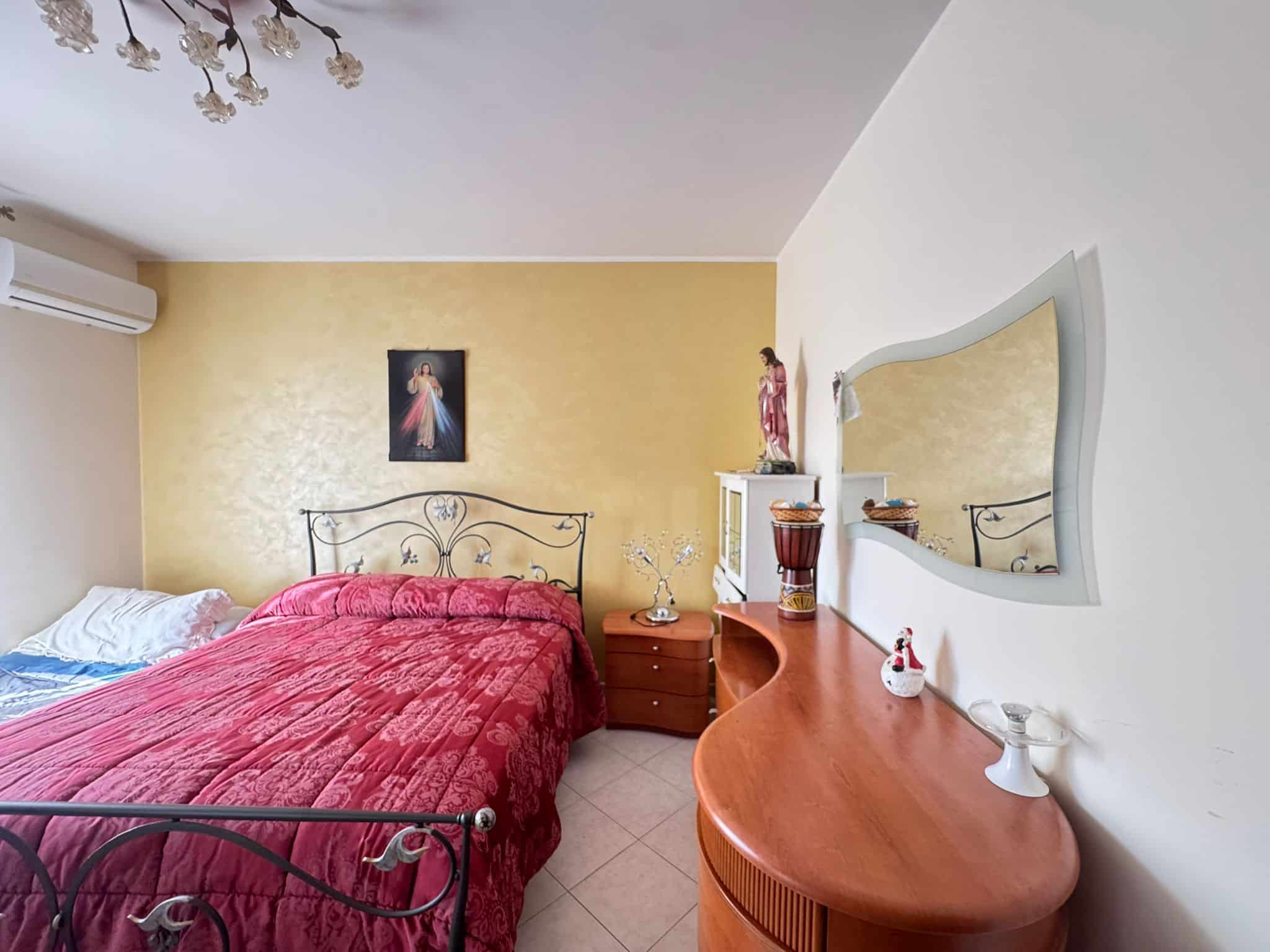Dettaglio di una camera da letto con letto a baldacchino in ferro battuto, pareti gialle e bianche, specchio a muro, decorazioni religiose e mobili in stile classico, luminoso e accogliente.