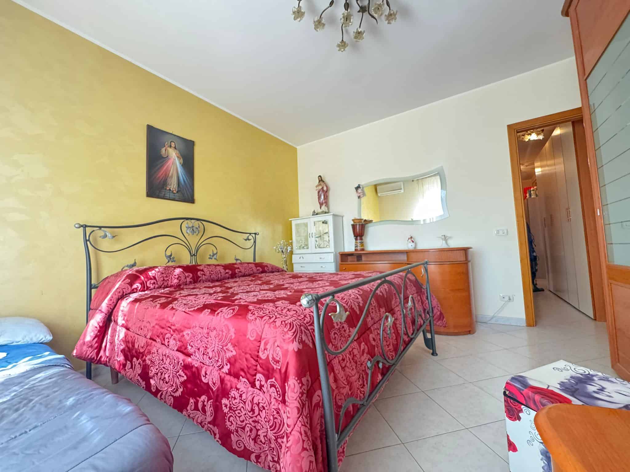 Letto matrimoniale con letto in ferro battuto e biancheria rossa, arredamento classico, pareti gialle e bianche, decorazioni religiose, specchio grande, mobile portaoggetti in legno, ambiente luminoso e accogliente.