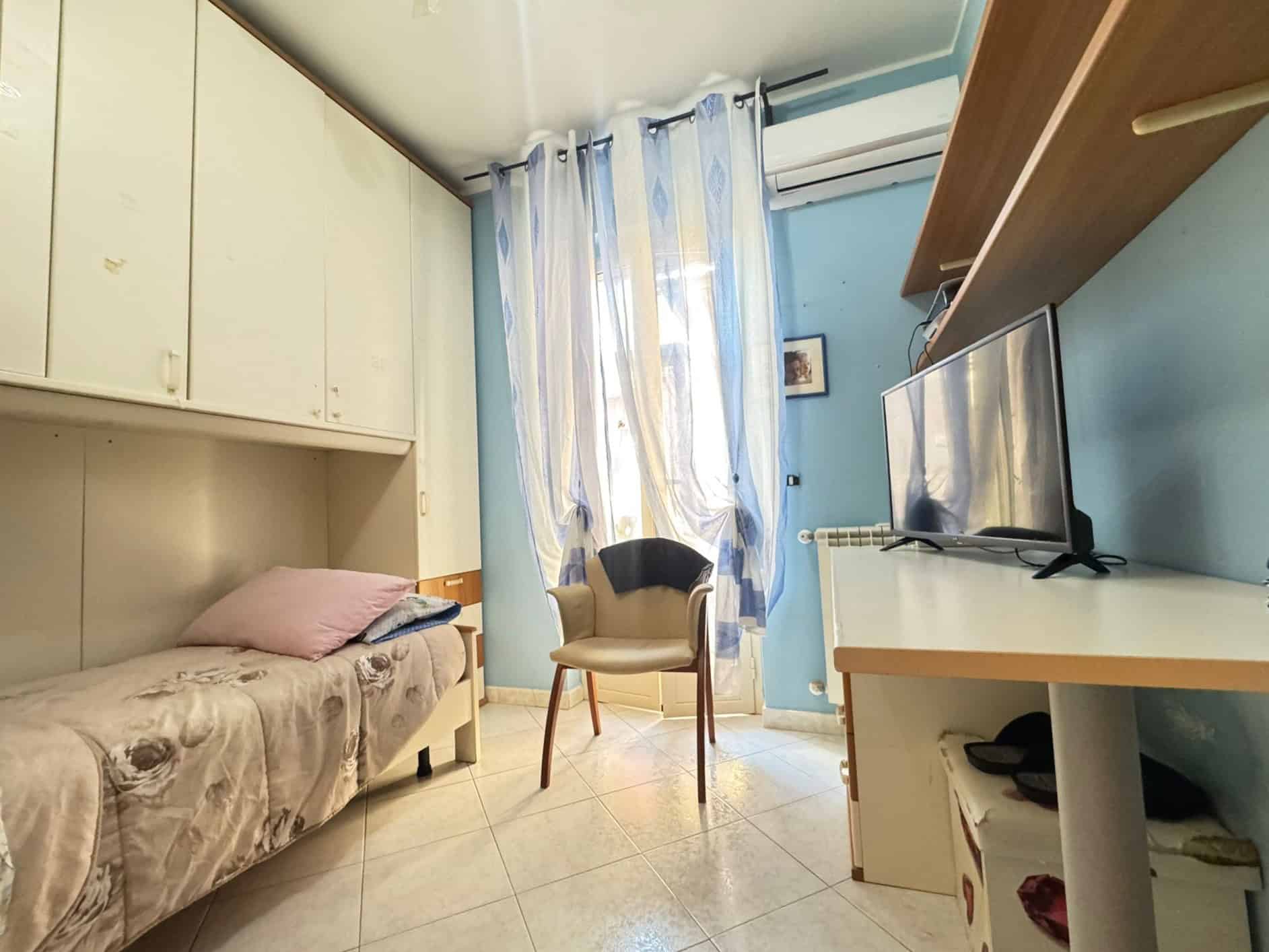 1. Divano singolo con cuscini in camera da letto vicino a finestra decorata con tende leggere blu e bianco, aria condizionata e televisione su mobile bianco, ambiente accogliente e luminoso.