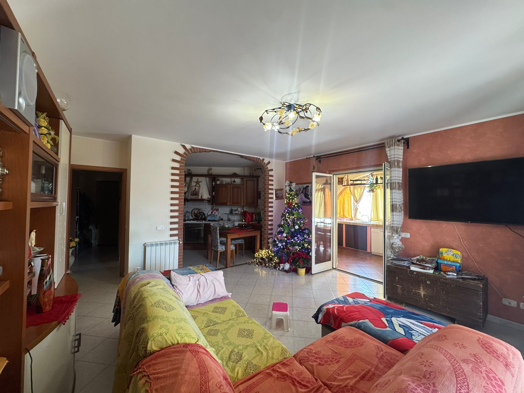 Ampio soggiorno con divani colorati, decorato natalizio con albero e luci, apertura sulla balconata soleggiata, cucina in stile rustico, ambiente caldo e accogliente, ideale per vivere momenti in famiglia e amici.