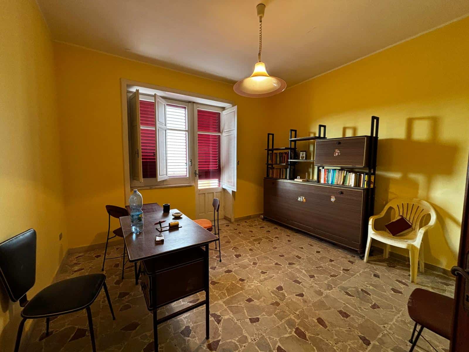 1. Interno soggiorno con pareti gialle, finestra con persiane rosse e mobili in stile classico, pavimento in graniglia, atmosfera luminosa e accogliente.