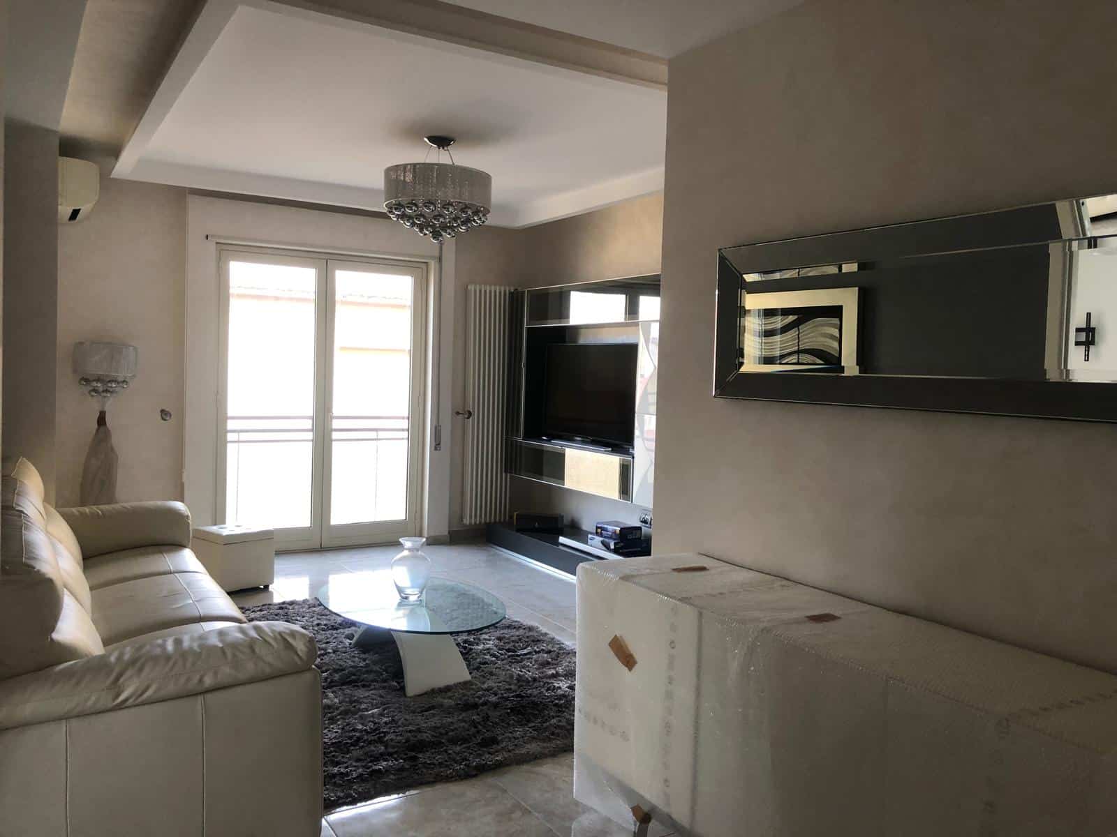 Immagine di un soggiorno moderno ed elegante con divano beige, televisore a schermo piatto, lampade da parete e un grande balcone con porte a doppia anta che permette molta luce naturale.