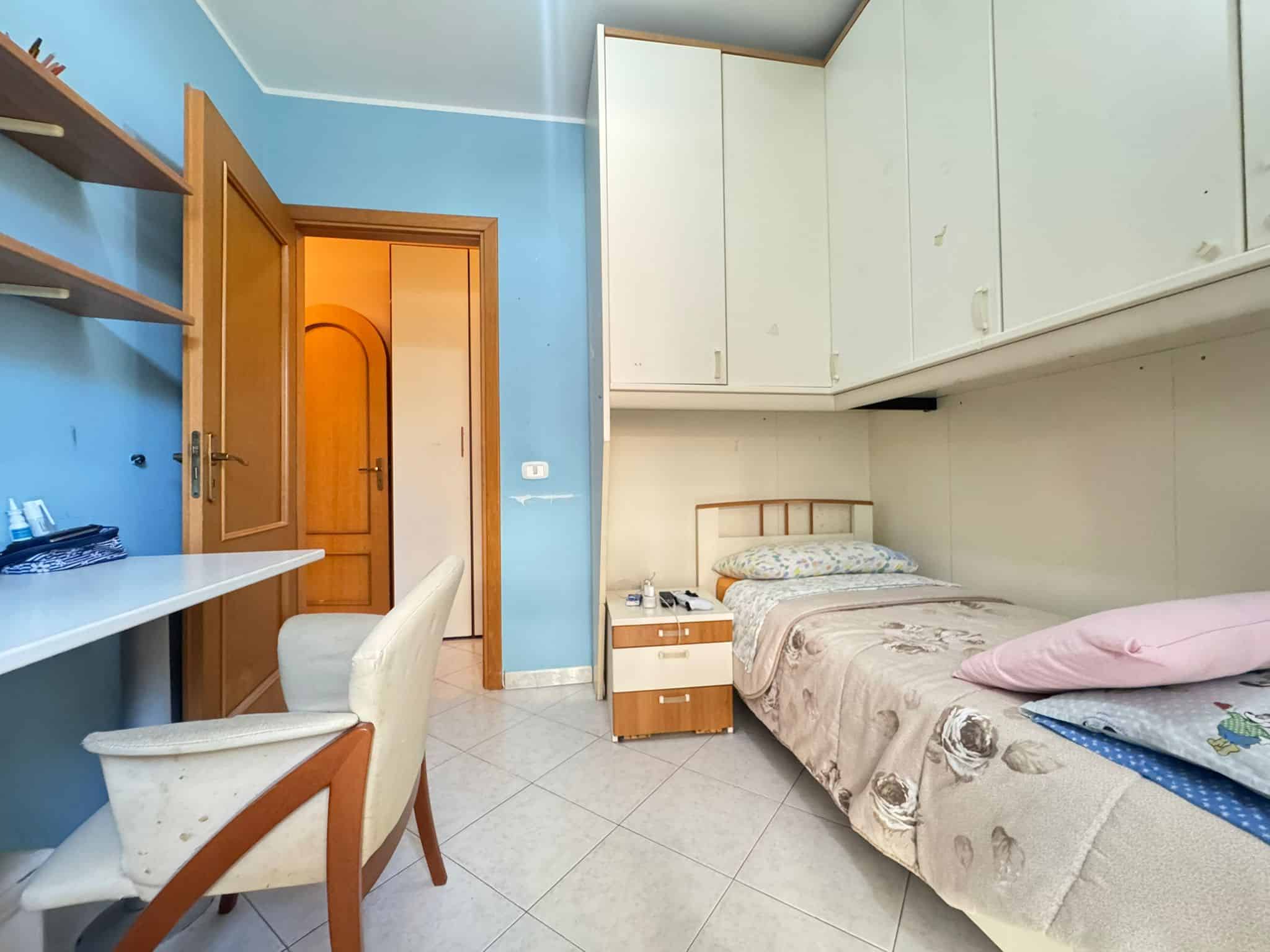 Pittura azzurra, camera da letto arredata, armadio a muro, letto singolo con biancheria, comodino con telefono e lampada, scrivania con sedia, porta di legno, ambiente abitabile in stile semplice.