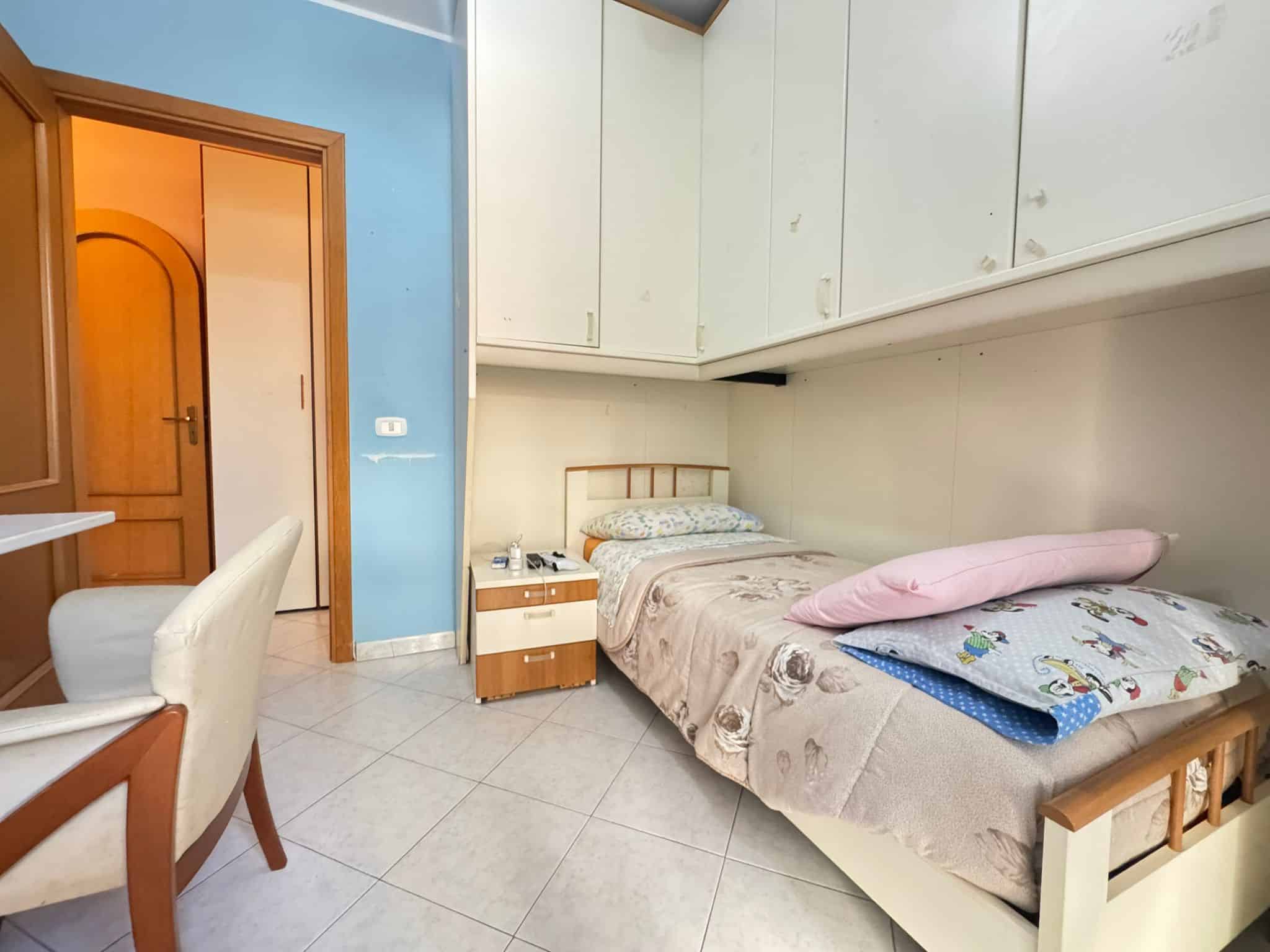 Letto singolo in una camera semplice con armadiature sopra il letto, lampada da tavolo e mobili funzionali, ideali per affitto o uso temporaneo.
