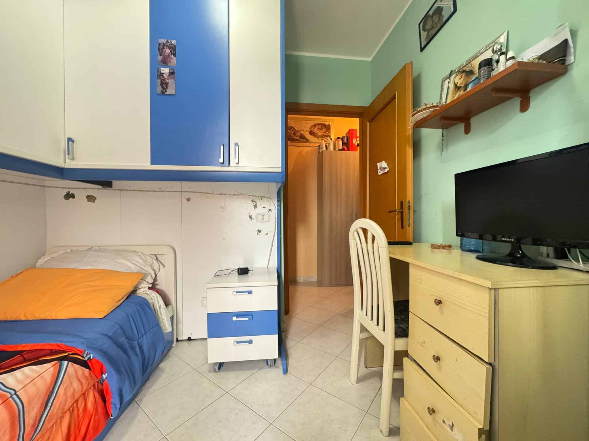Letto singolo con cuscino arancione, armadio a muro blu e bianco, scrivania con TV, pareti verde, porta in legno, scaffale con oggetti e decorazioni, ambiente abitativo funzionale.