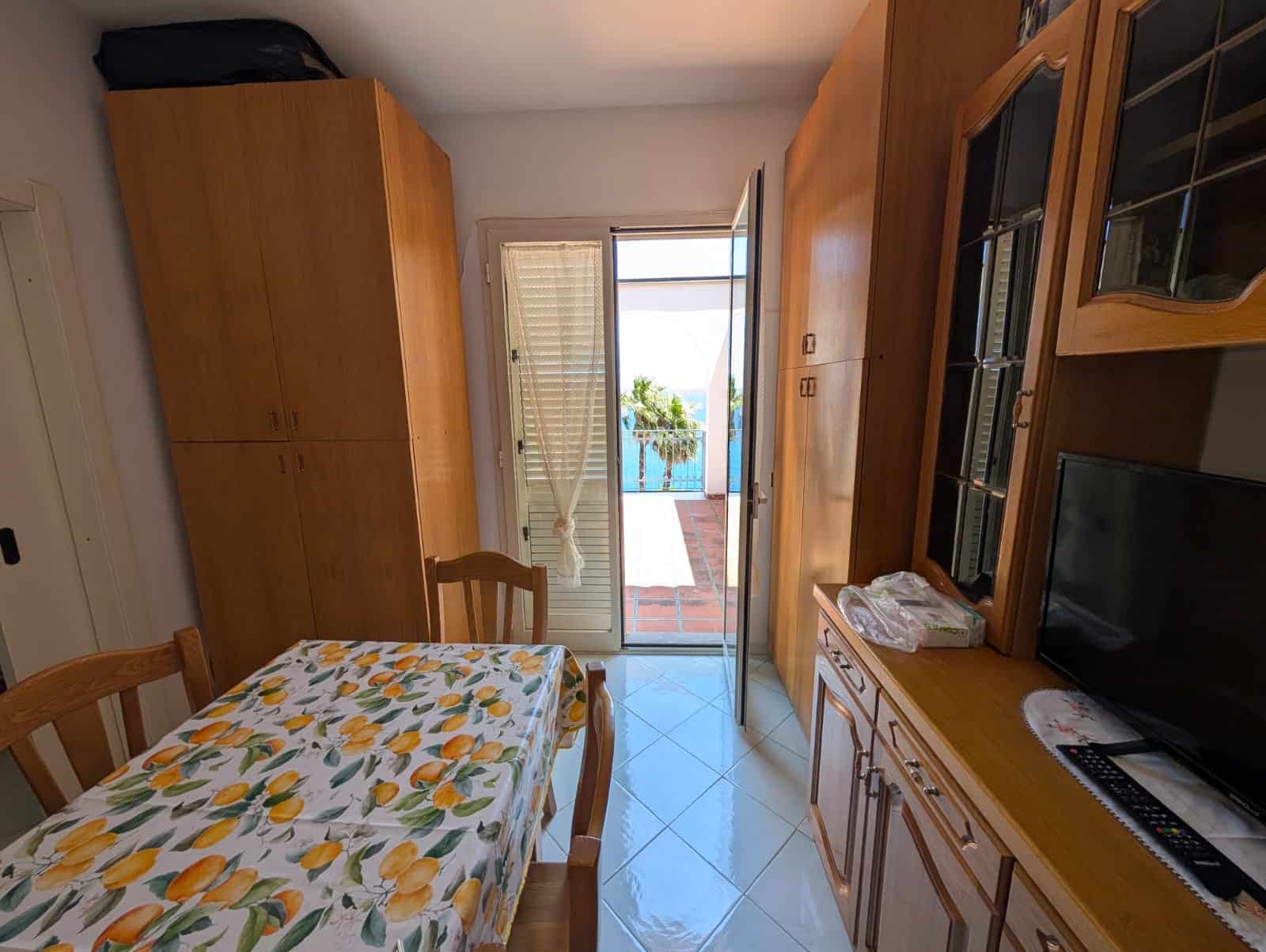 Affitto appartamento con balcone, vista mare, arredi in legno, soggiorno luminoso, zona pranzo, TV, tavolo con tovaglia di limoni, spazio esterno con vista panoramica.
