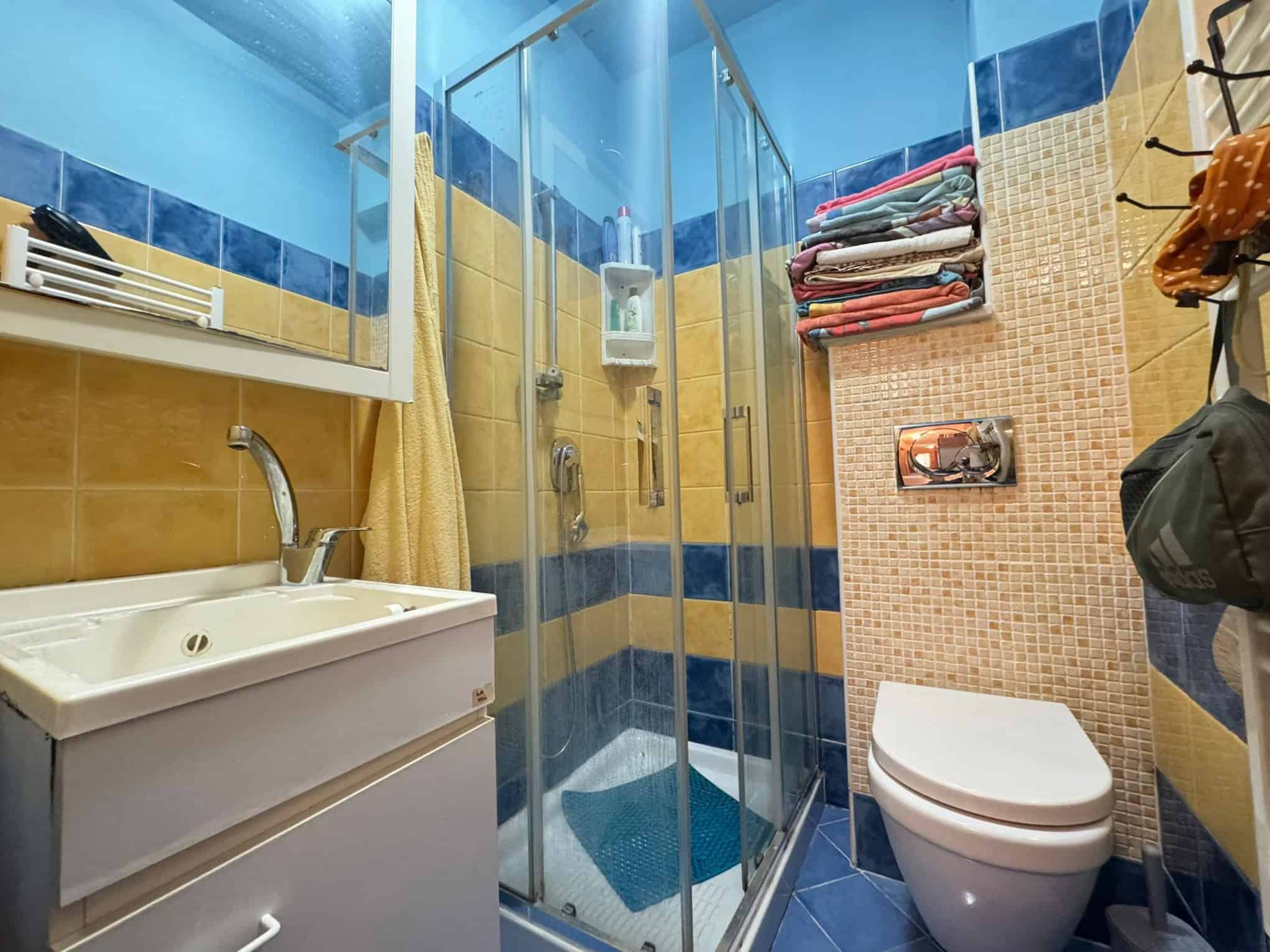 Ampia doccia in bagno con pareti in vetro, sanitari moderni, rivestimenti colorati, asciugamani piegati su mensola, dettagli di arredo utili e pratici, spazio ottimizzato e design funzionale.