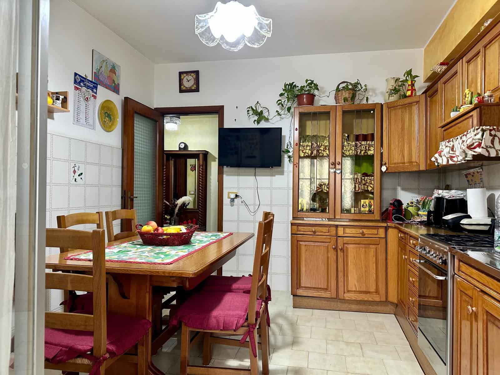Cucina classica con mobili in legno, tavolo da pranzo con tovaglia e sedie con cuscini rossi, pareti rivestite di piastrelle bianche, elettrodomestici e decorazioni, spazio accogliente e funzionale.