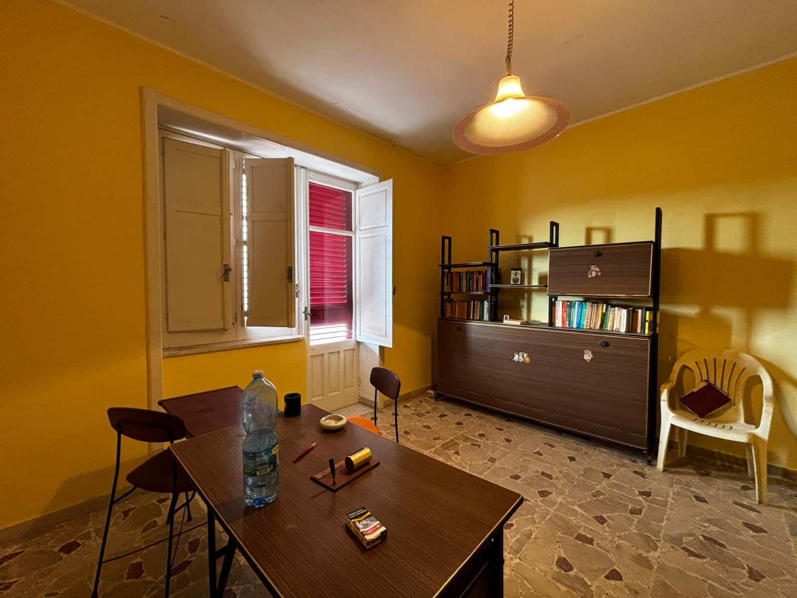 Appartamento in affitto a Napoli, ampio soggiorno con pareti gialle, finestra con persiana rossa, comoda scrivania, libreria moderna, pavimento in mosaico, atmosfera luminosa e accogliente.