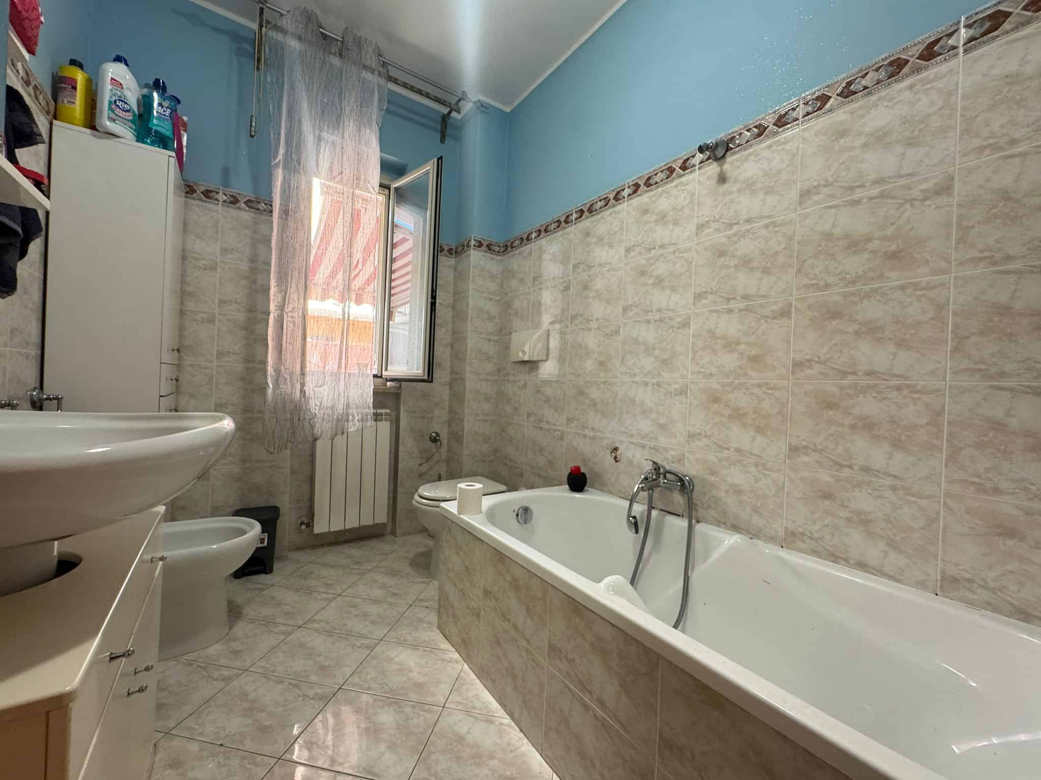 Bagno con vasca, bidet, lavabo e finestra aperta, con pareti e pavimenti in piastrelle beige, ideale per soluzioni immobiliari e affitti a NextCasa.
