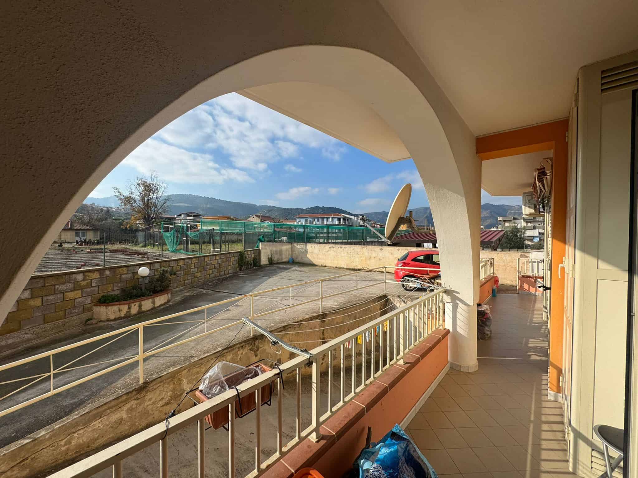 Balcone soleggiato con vista su cortile, montagna e cielo azzurro, arredato con piante e oggetti vari, in zona residenziale tranquilla, con strada asfaltata e parcheggio.