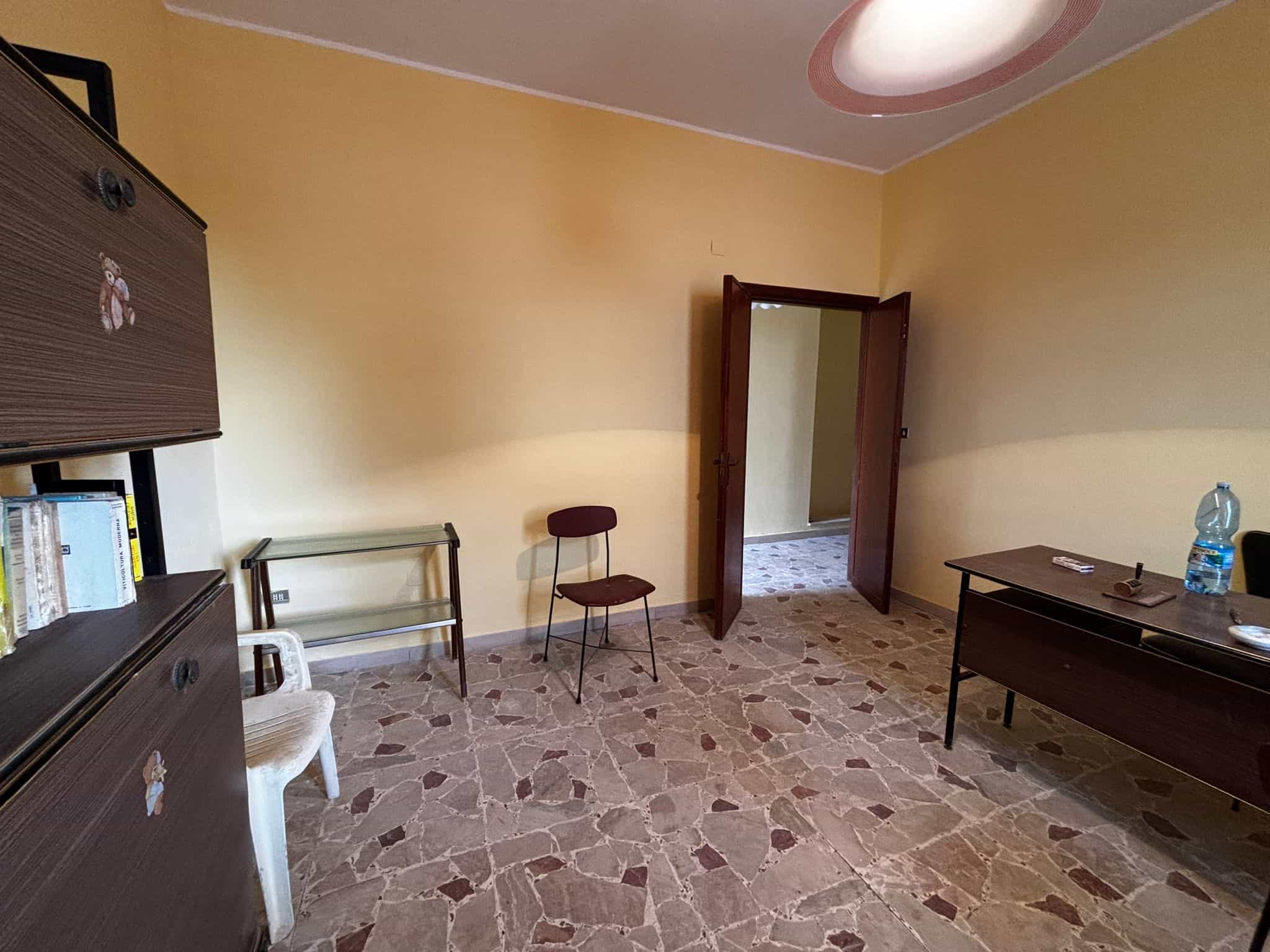Poltrona di plastica bianca e sedia imbottita in tessuto marrone, mobili in legno scuro, pareti gialle, pavimento in piastrelle di marmo e porta in legno aperta verso un ingresso in un appartamento.