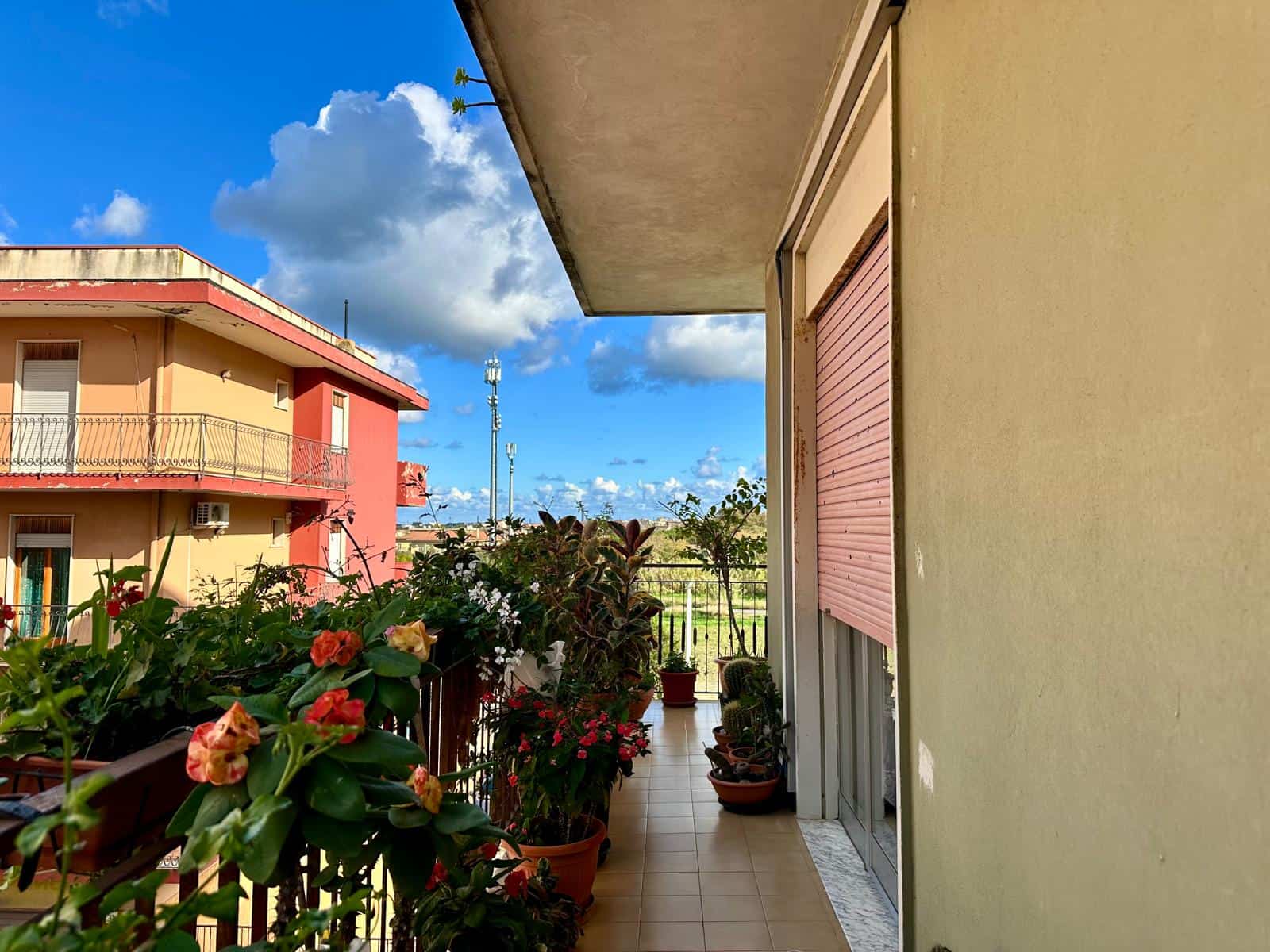 Balcone in un appartamento residenziale con piante e fiori, vista su edificio colorato e cielo blu con nuvole, ambiente luminoso e accogliente, ideale per relax e cura delle piante.