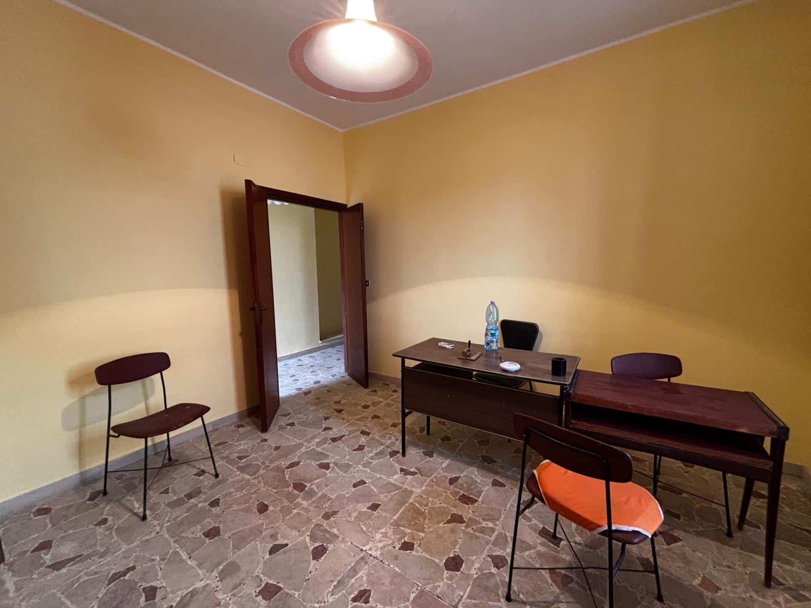 Affaccio sulla stanza vuota di ufficio con pareti gialle, scrivania in legno, sedie, bottiglia d'acqua e oggetti vari, ambiente luminoso e semplice, ideale per lavoro o attività professionali.