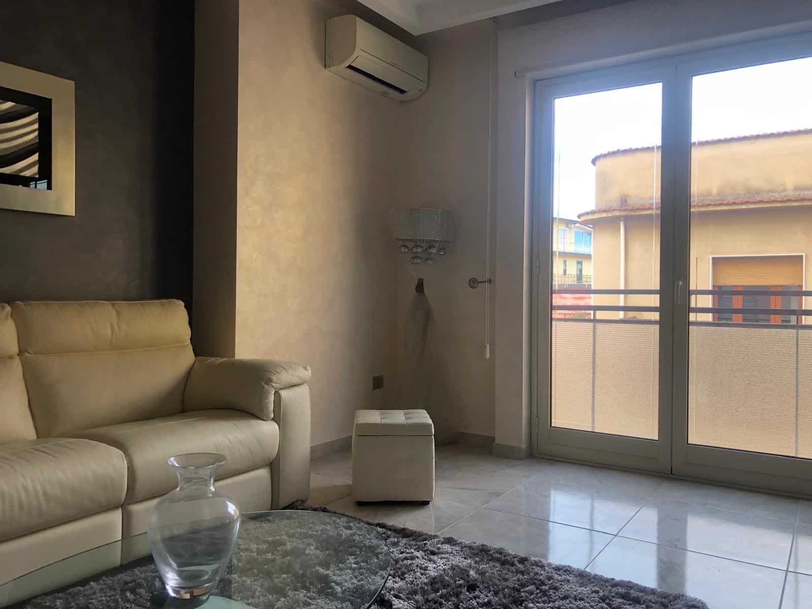 Soggiorno moderno con divano in pelle beige, grande terrazza con vista su edificio urbano, aria condizionata e arredamento elegante, ideale per case in vendita o affitto a NextCasa.