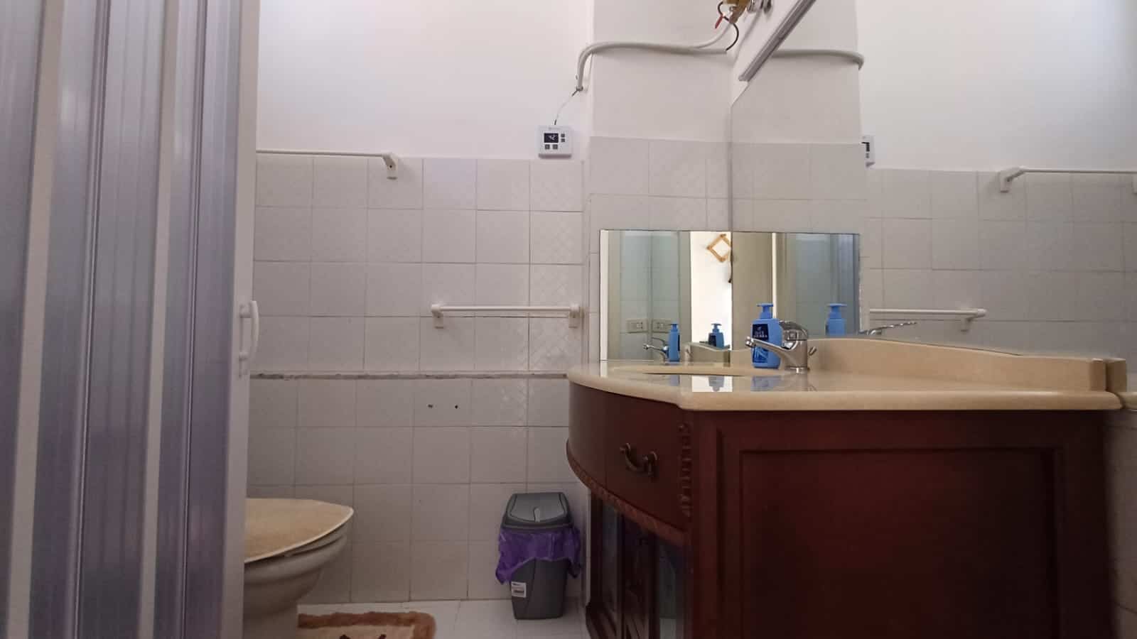 Lavabo bagno con mobile in legno, specchio, dispenser di sapone e asciugamano in bagno di servizio. La stanza presenta piastrelle bianche e vari accessori utili per l'igiene quotidiana.