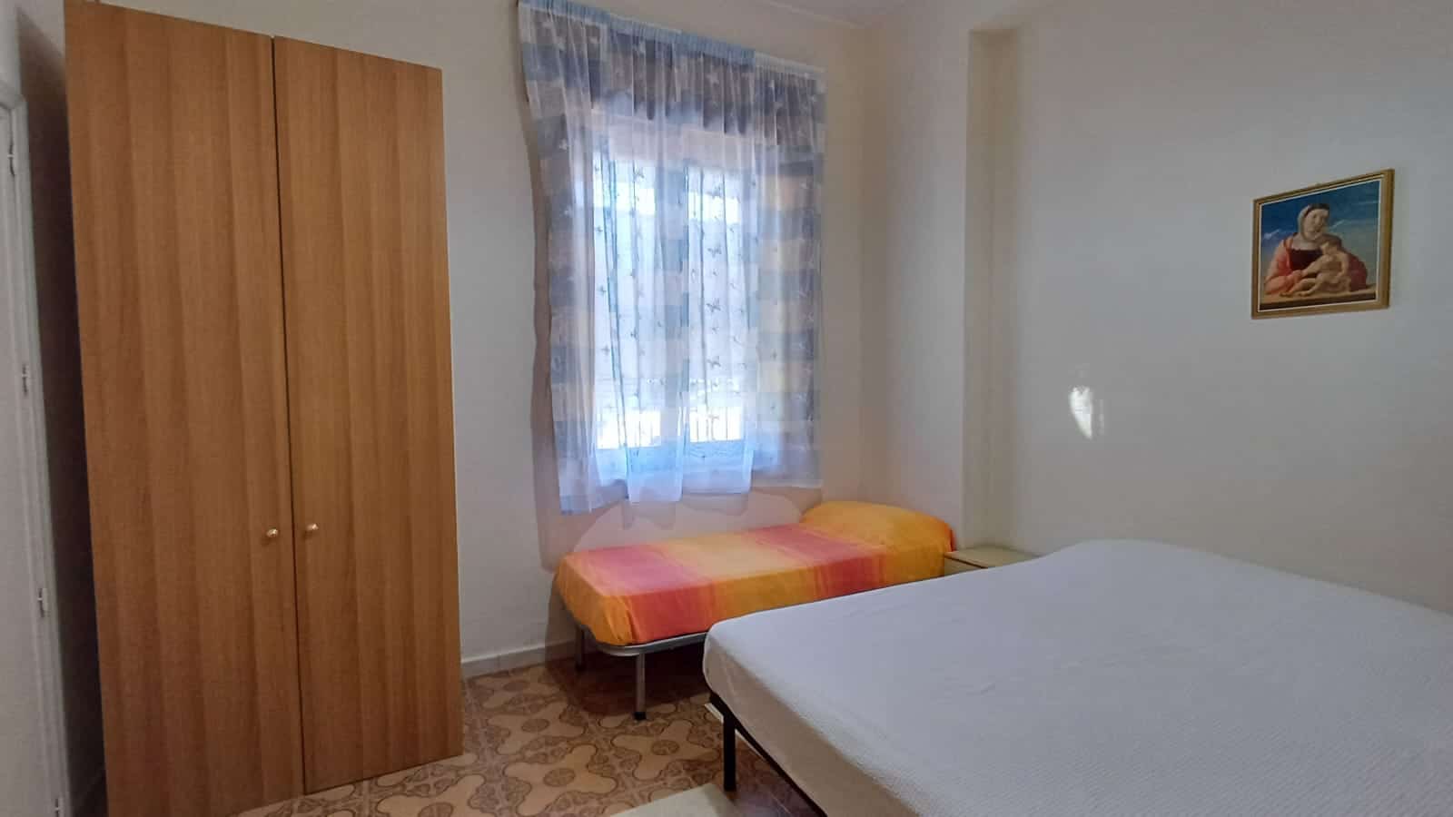 Camere da letto in affitto con arredi semplici, letto matrimoniale e singolo, finestra con tende leggere, ambiente luminoso e accogliente, ideale per soggiorni temporanei o affitti brevi.
