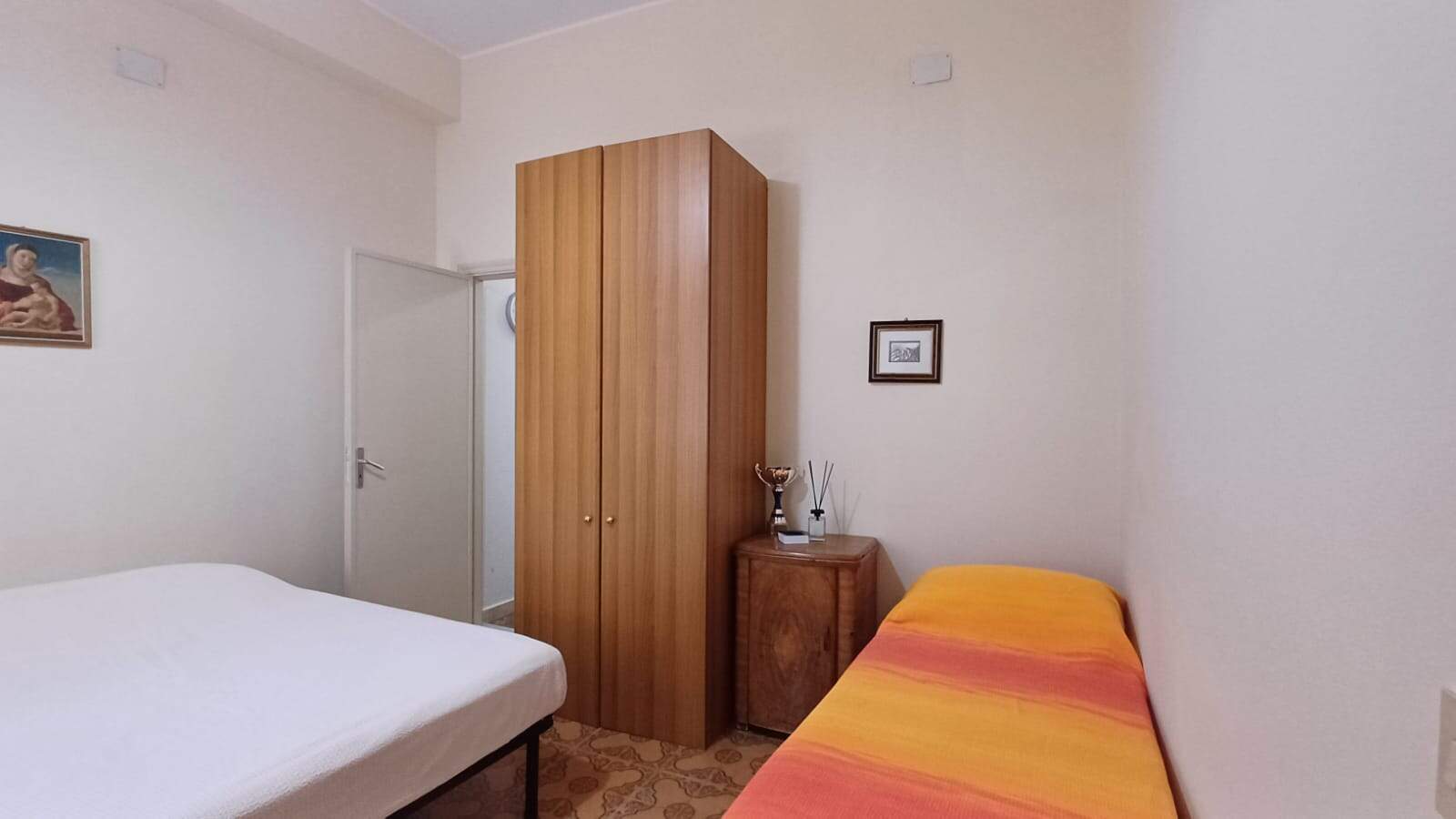 Stanza da letto semplice con armadio in legno, letto singolo con copripiumino colorato e decorazioni minimaliste in ambiente luminoso e accogliente.