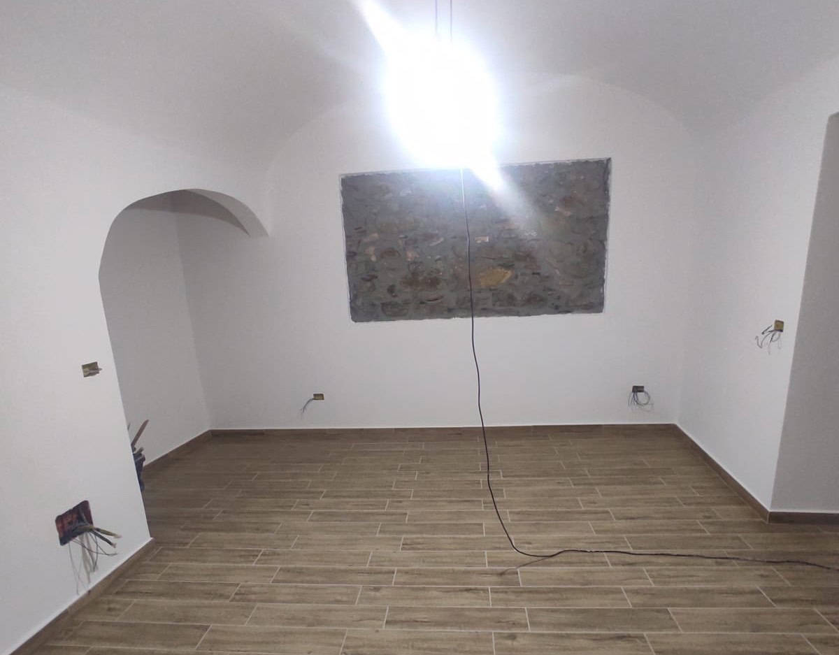 Ripresa di una stanza in fase di ristrutturazione con pareti bianche, pavimento in gres effetto legno e una parete in pietra dietro la finestra, pronto per arredare.